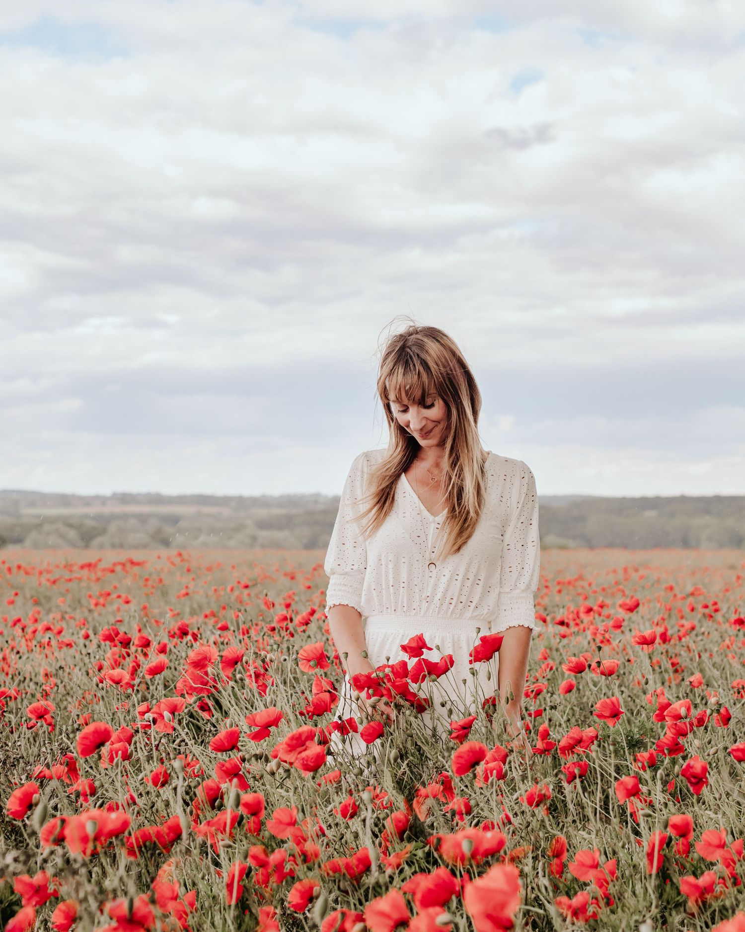 A woman standing in a vibrant field of red poppies under an overcast sky.