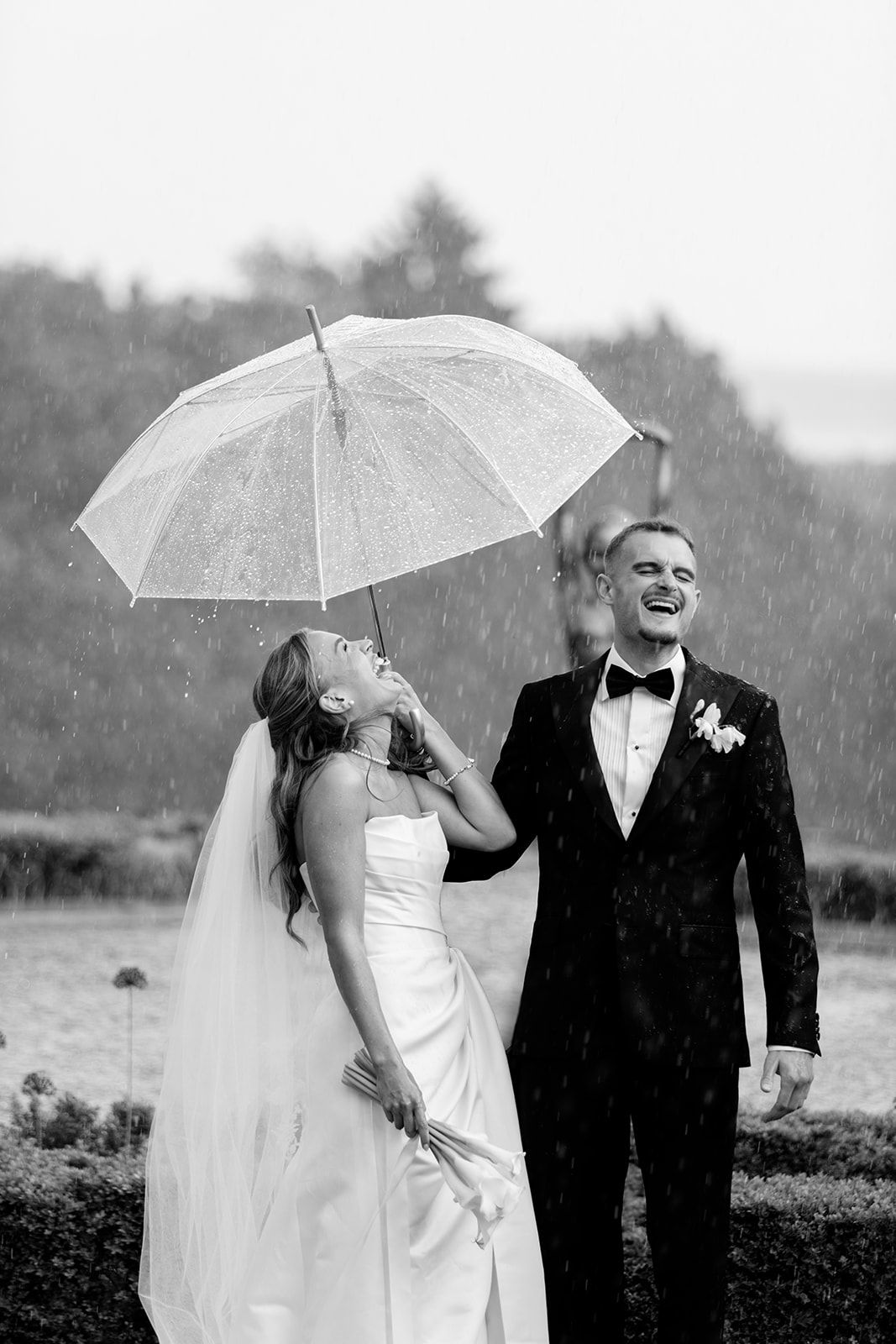 A couple shares a joyful moment under a white umbrella in the rain during their wedding celebration.