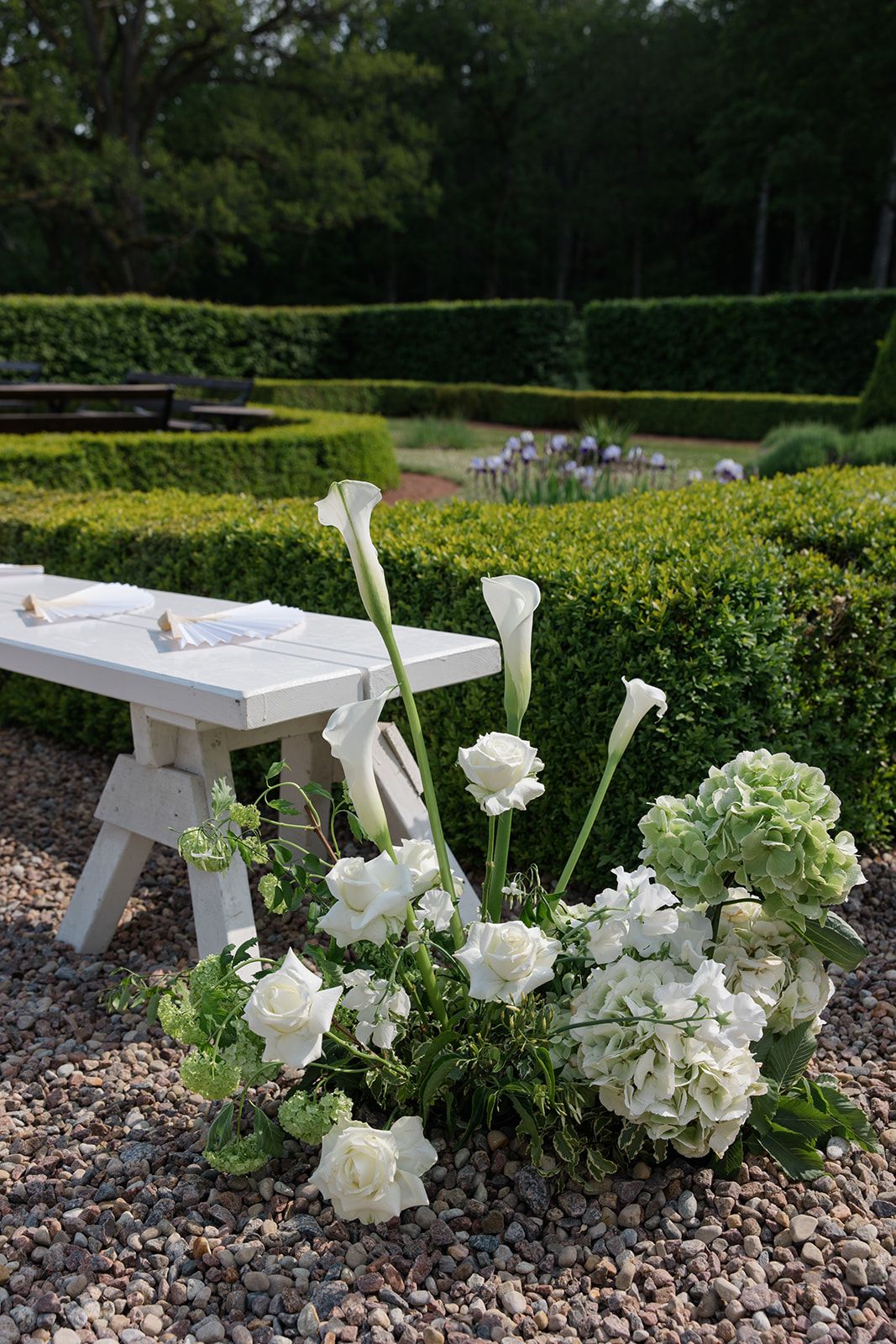 White roses elegantly arranged on a wooden bench in a formal garden with manicured hedges and gravel path.