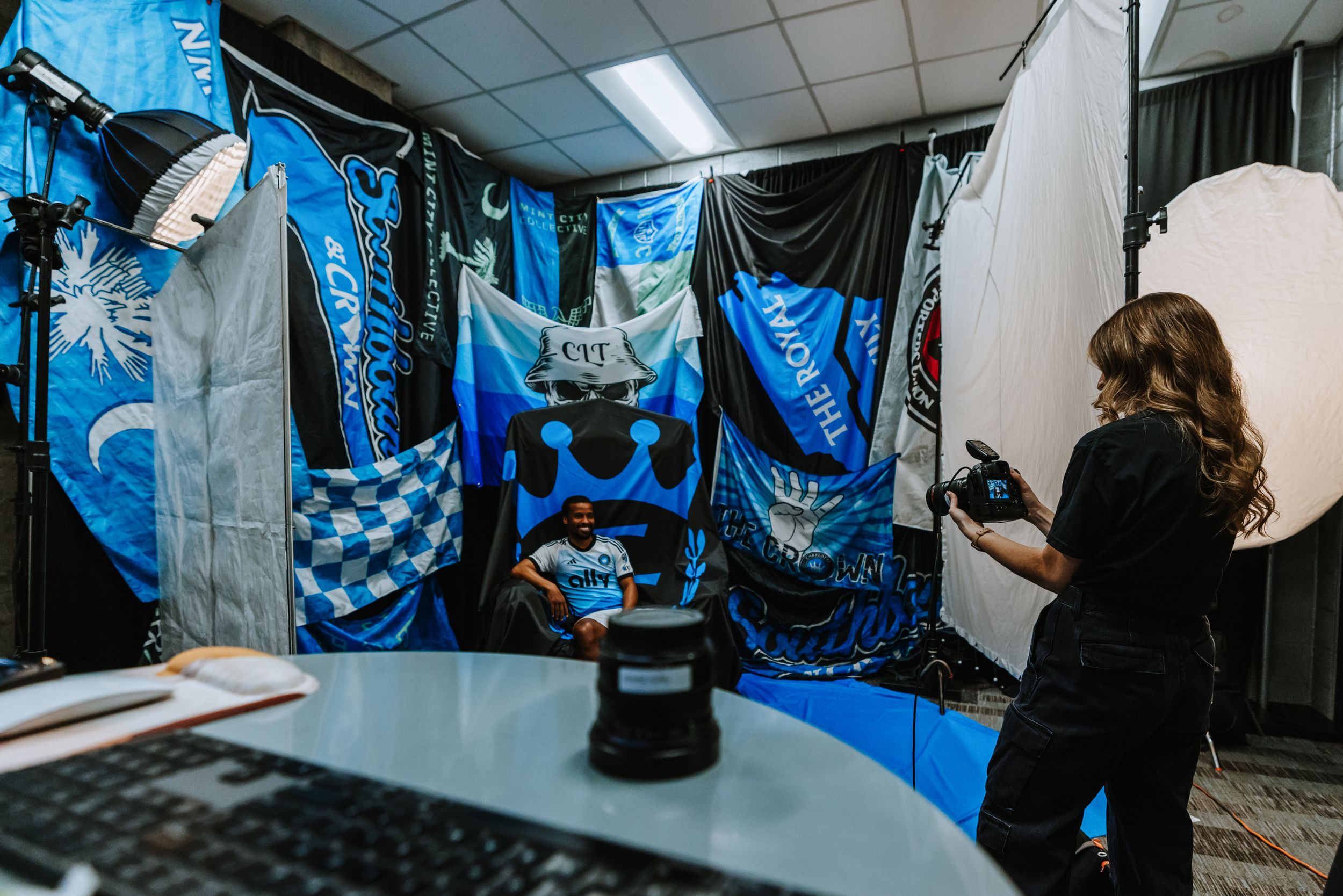 A female photographer photographs a Charlotte FC player seated before a wall of blue supporter flags.