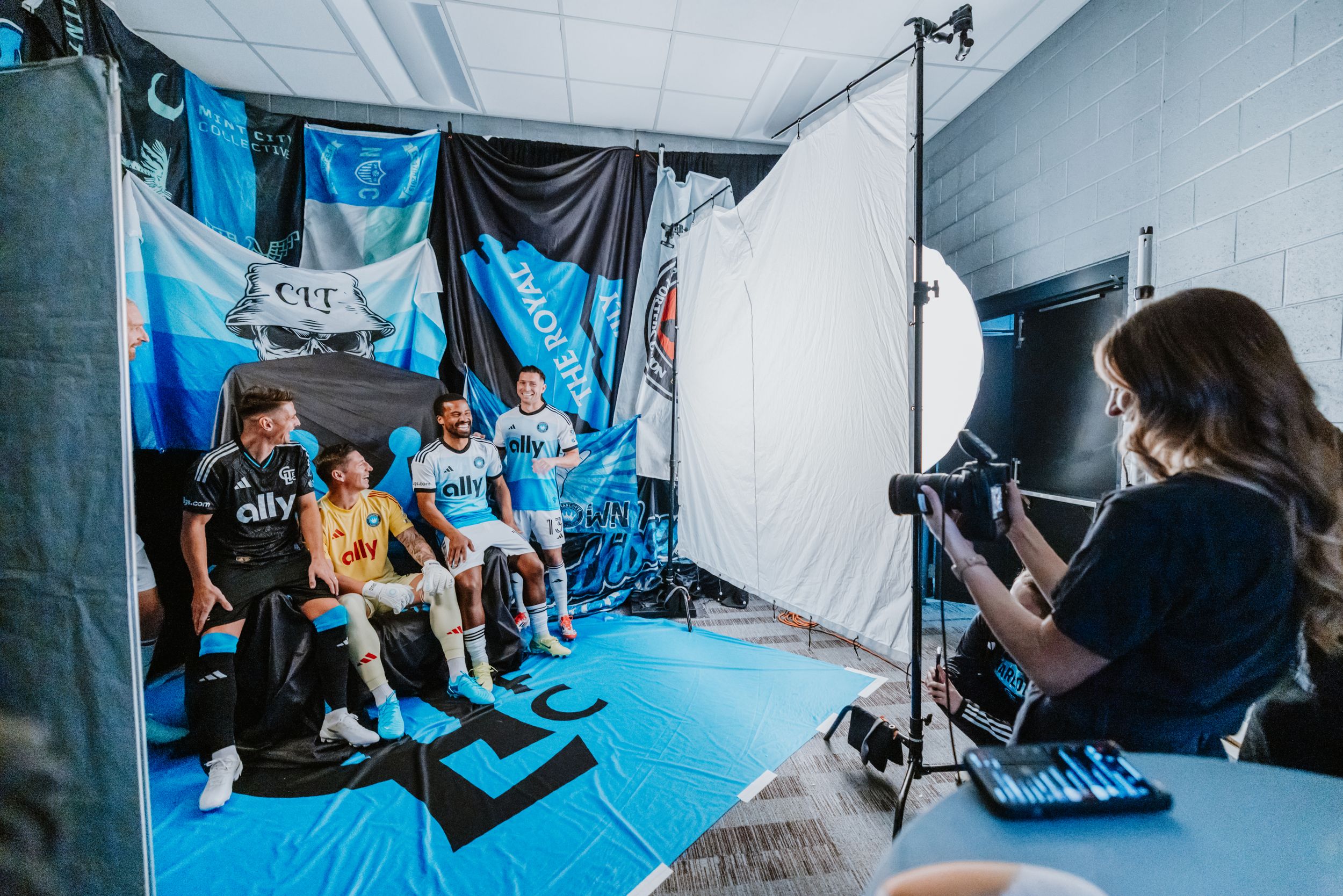 A female photographer shoots Charlotte FC players in team jerseys against a backdrop of supporter flags.