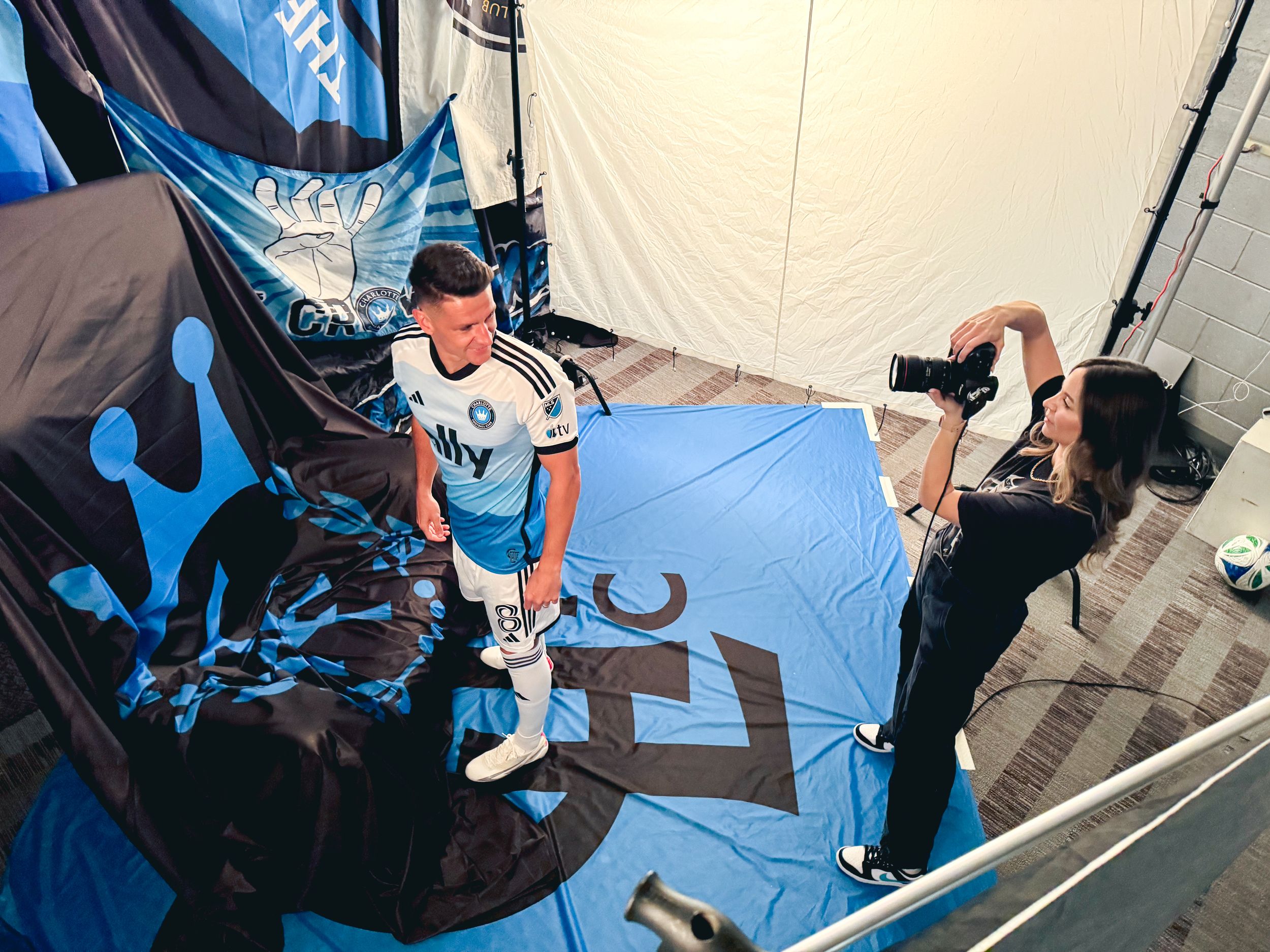 A female photographer shoots a Charlotte FC player in white jersey against blue supporter flag backdrop.