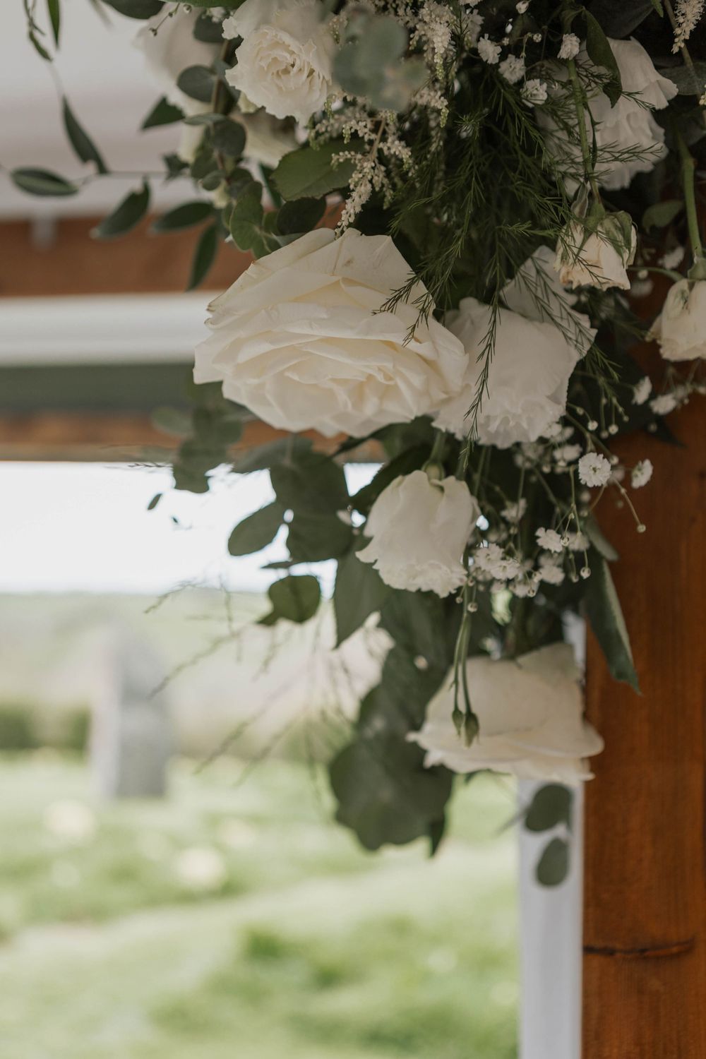 Close-up of white roses and eucalyptus floral arrangement on rustic wooden wedding arch.
