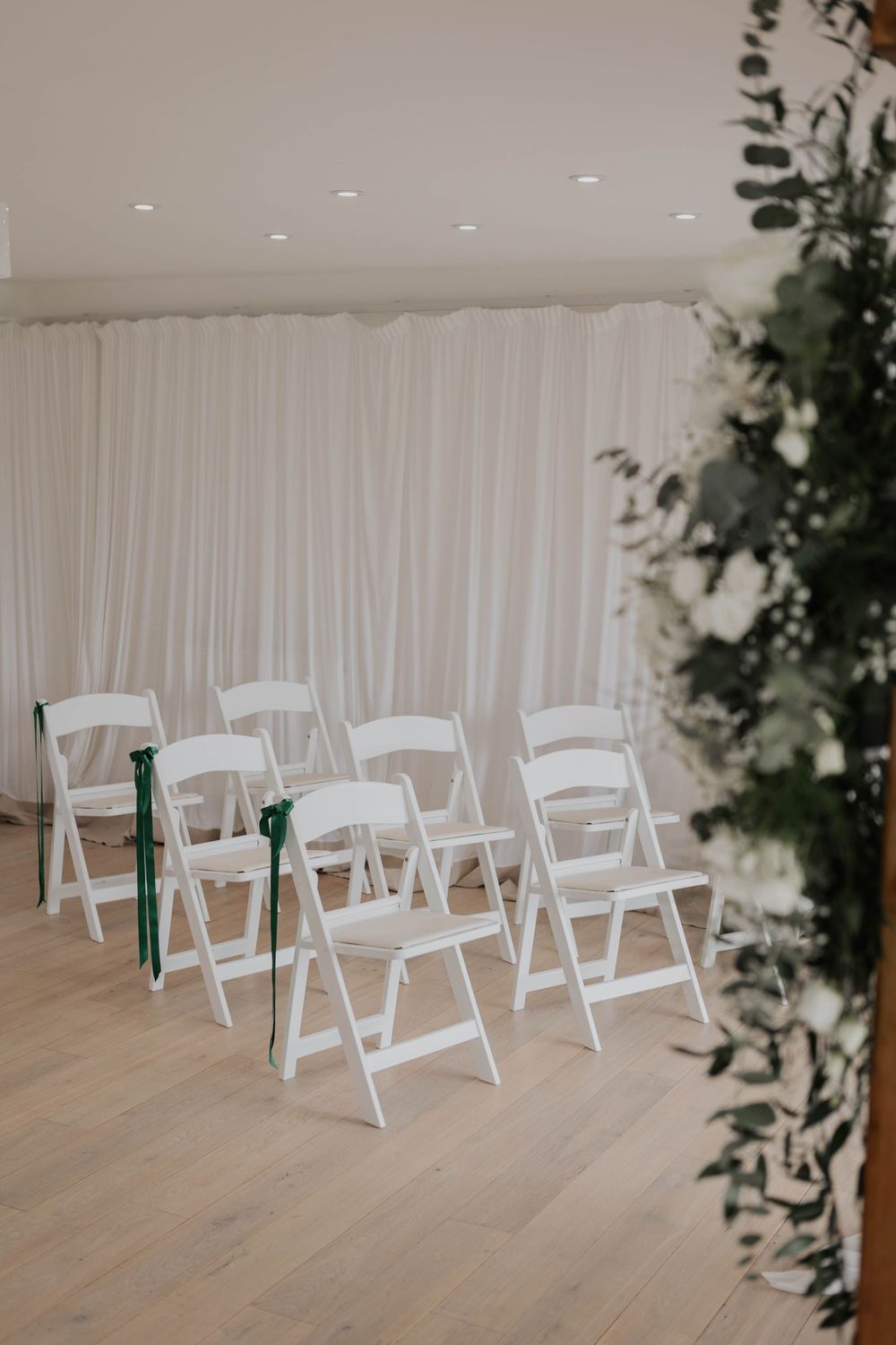 Rows of white folding chairs with green ribbon bows in bright wedding ceremony space.