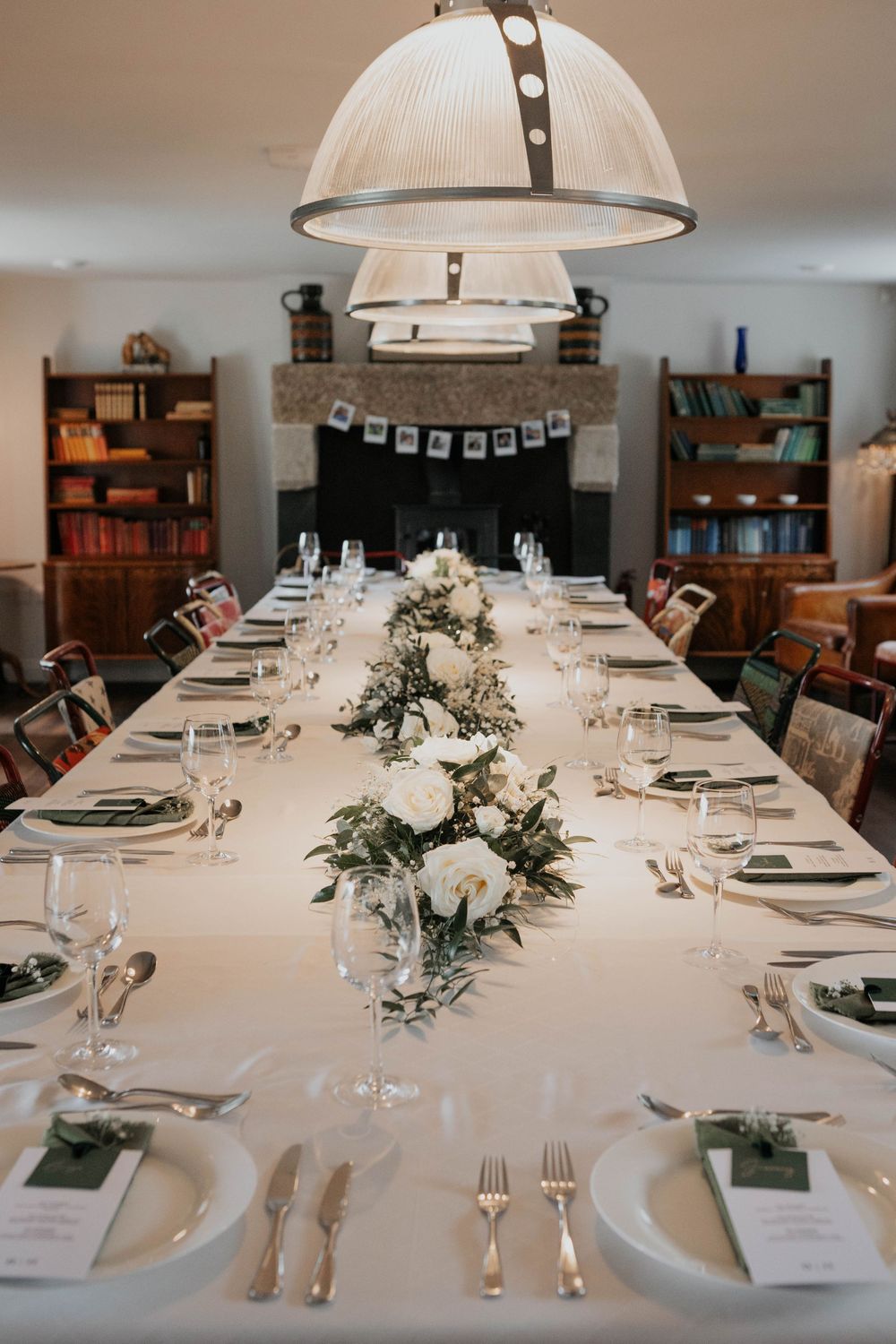 Elegant long dining table set with white florals, greenery, glassware, and place settings under pendant lights.