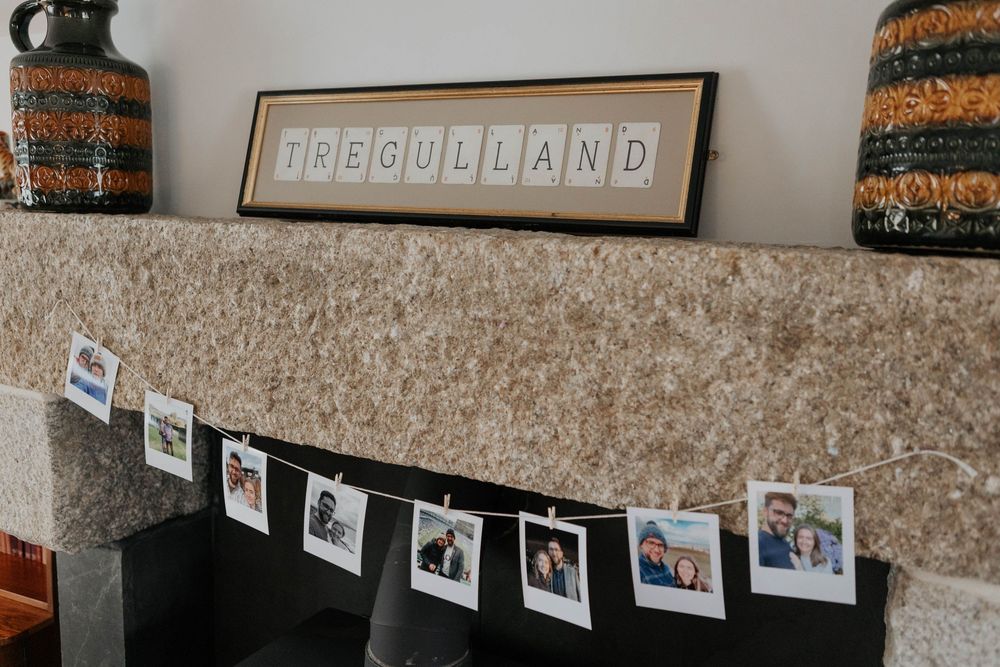 Scrabble letter sign spelling TREGULLAND on mantelpiece with polaroid photo garland hung below on stone fireplace.