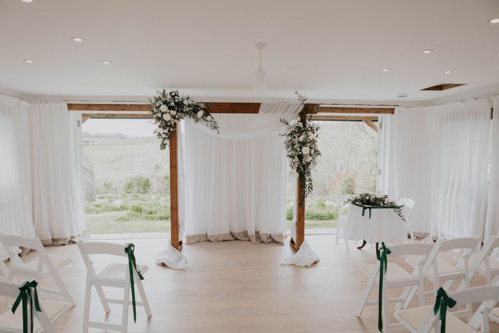White wedding ceremony room with wooden arch, floral decor and countryside window views.