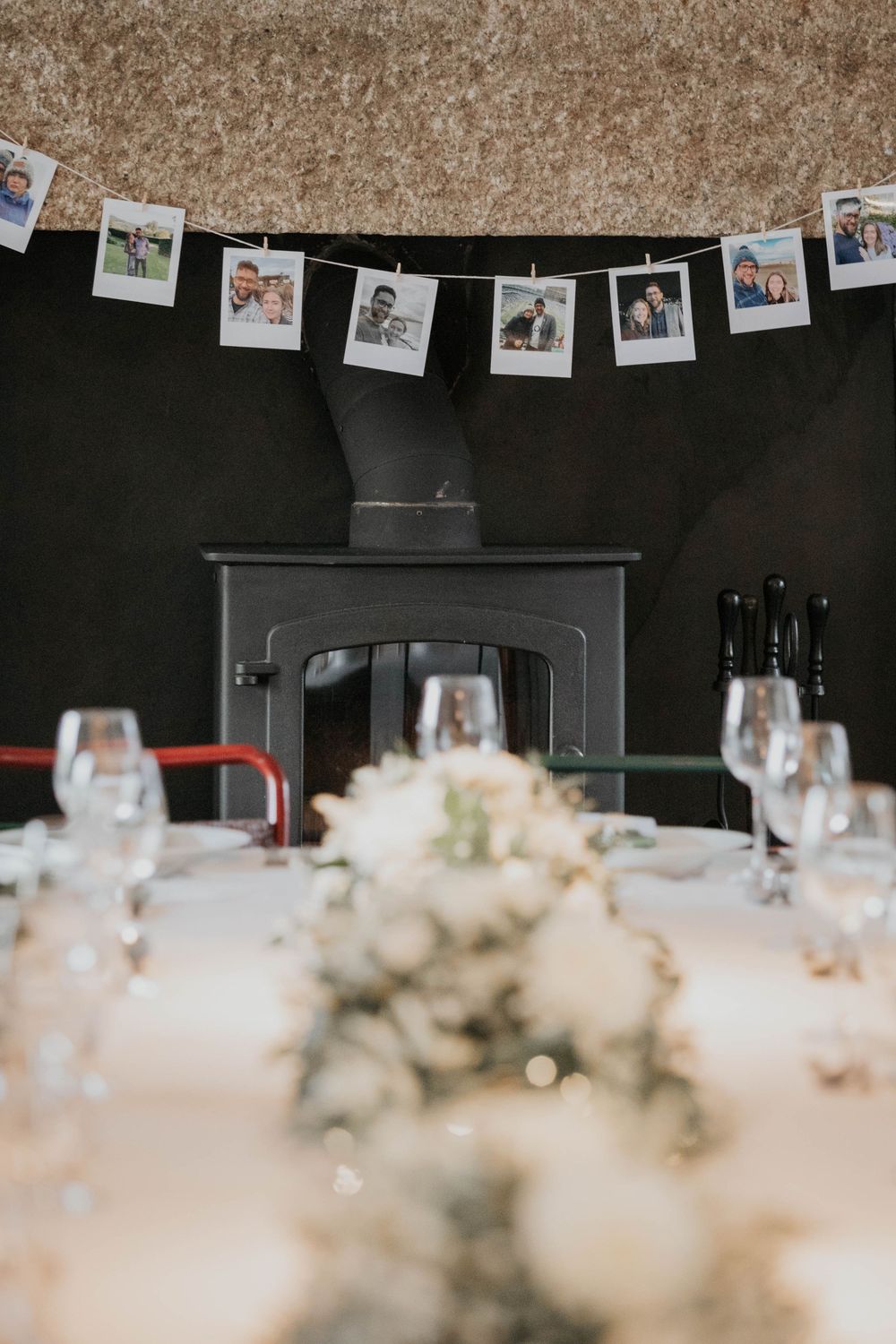 Intimate wedding reception table with white floral centerpiece and polaroid photo garland hanging above a black wood-burning stove.