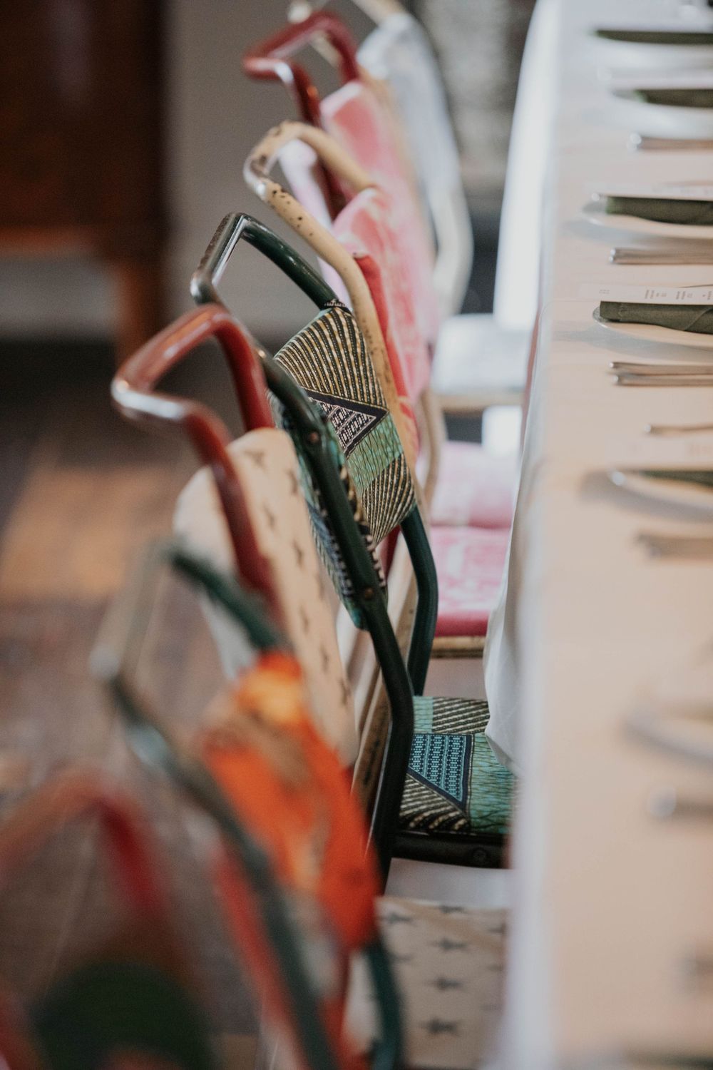 Row of eclectic mismatched colorful upholstered dining chairs lined up along a white wedding reception table.