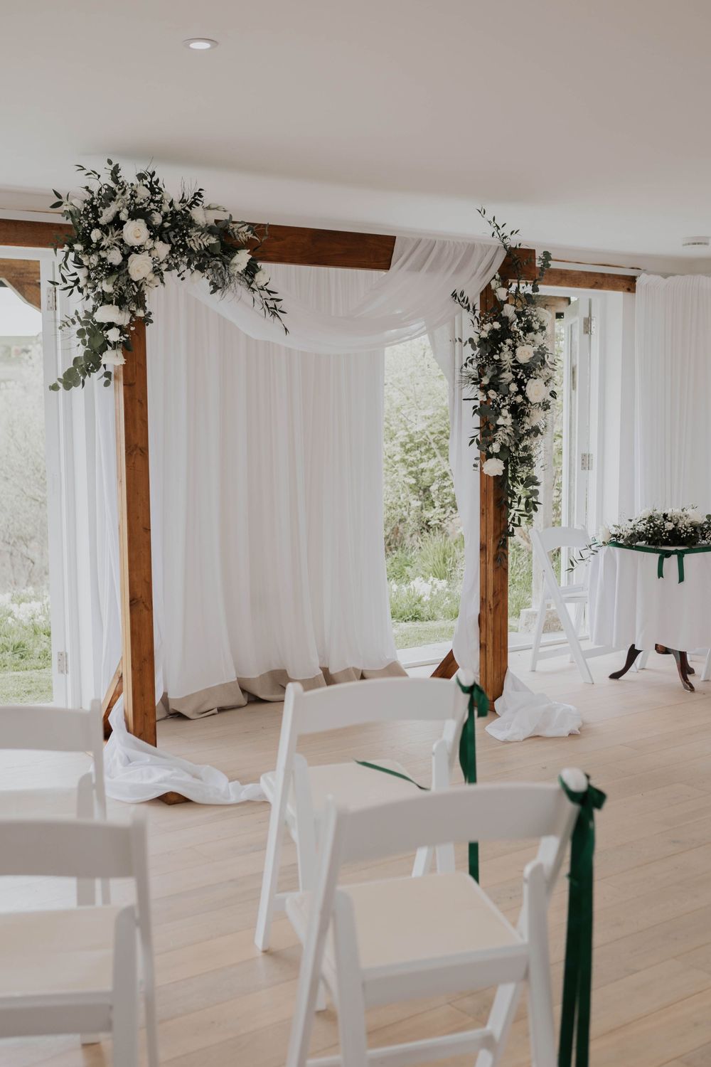 Rustic wooden arch with white draping and floral arrangements in bright ceremony room.