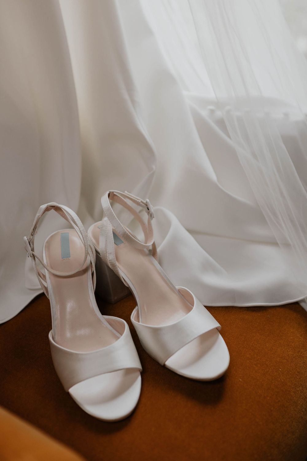 Elegant white satin block heel bridal sandals with ankle straps placed on an orange velvet chair.