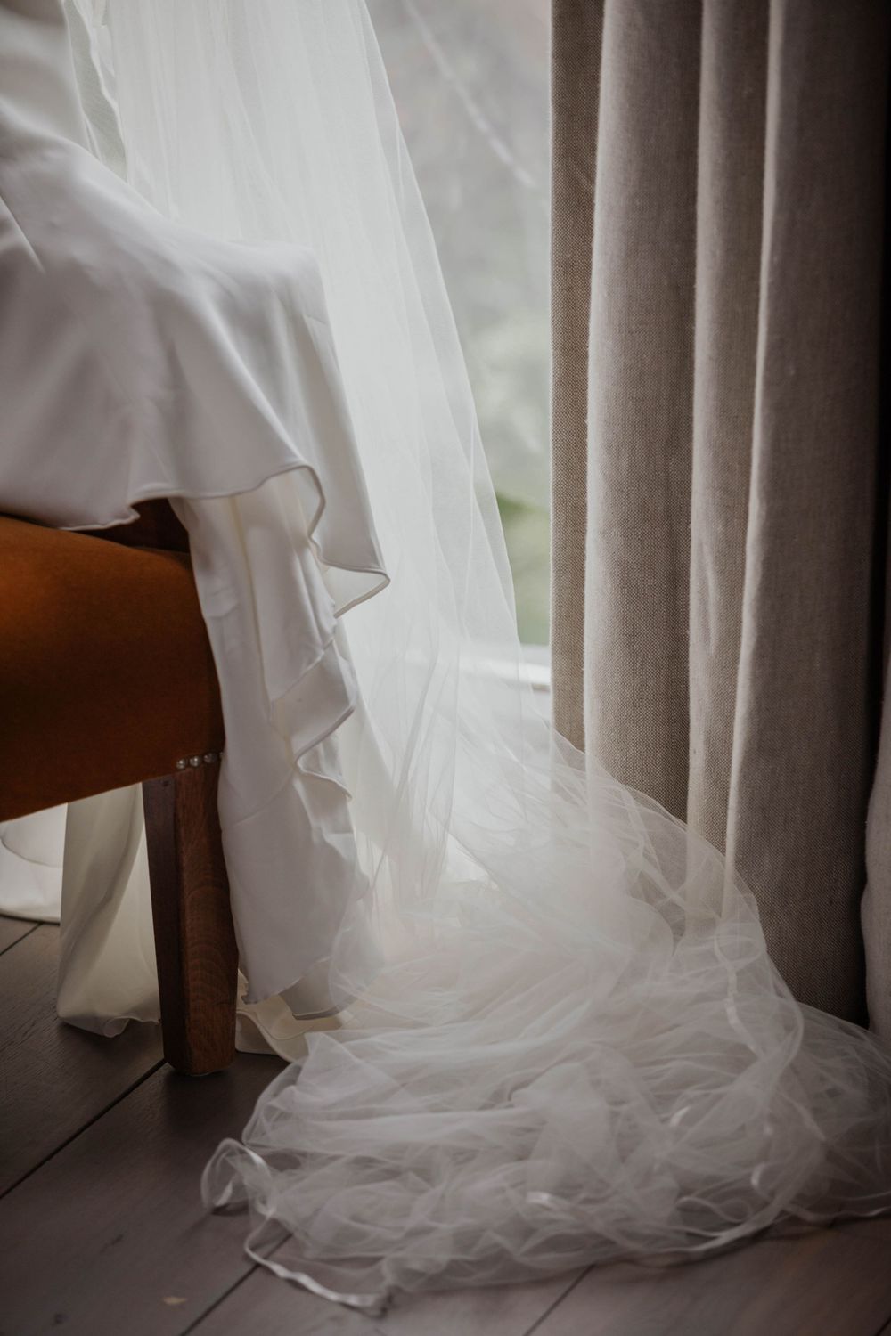 Elegant white wedding dress with tulle veil draped over wooden chair by window.