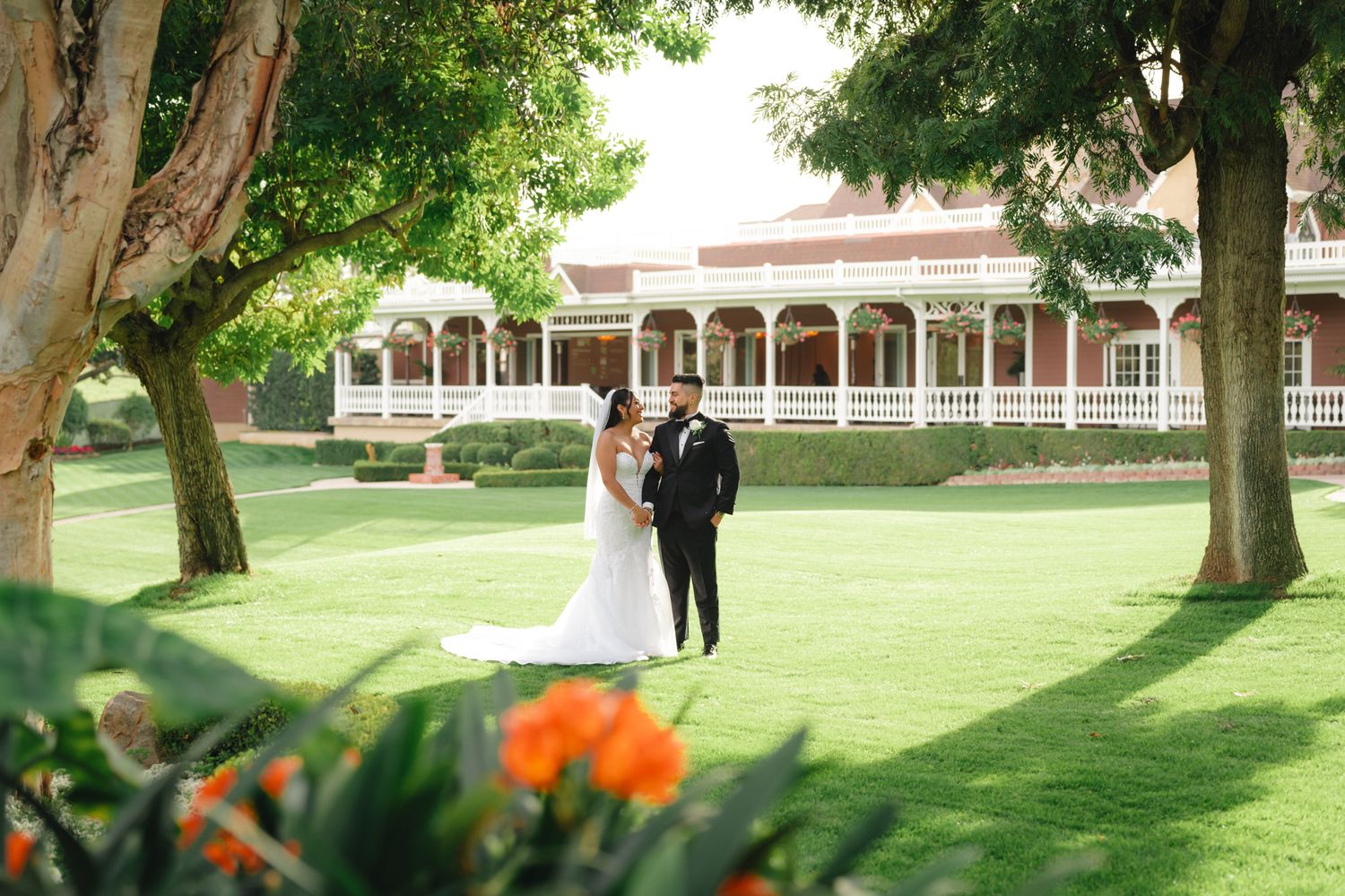 A couple walks together on a lush green lawn surrounded by mature trees on a bright sunny day.