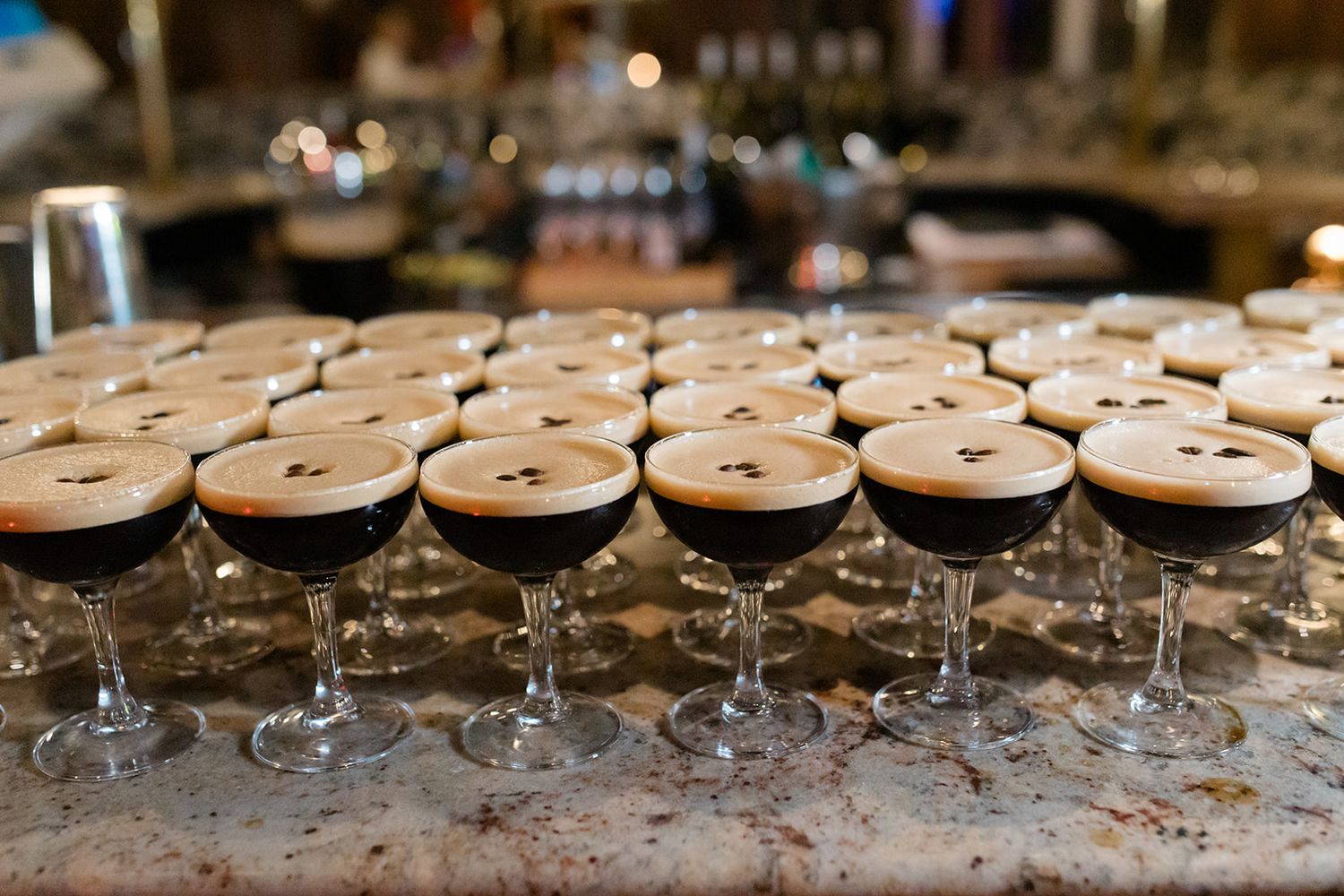 Rows of elegant cocktail glasses filled with espresso martinis line up on a dark bar counter at an event.