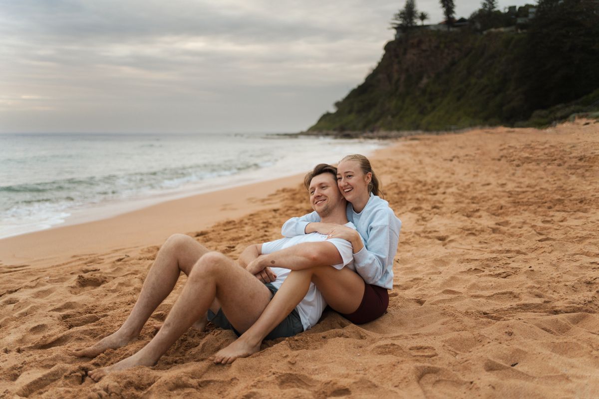 A couple sits together on a sandy Newport beach at sunrise with cliffs and ocean waves in the background during engagement photoshoot Sydney