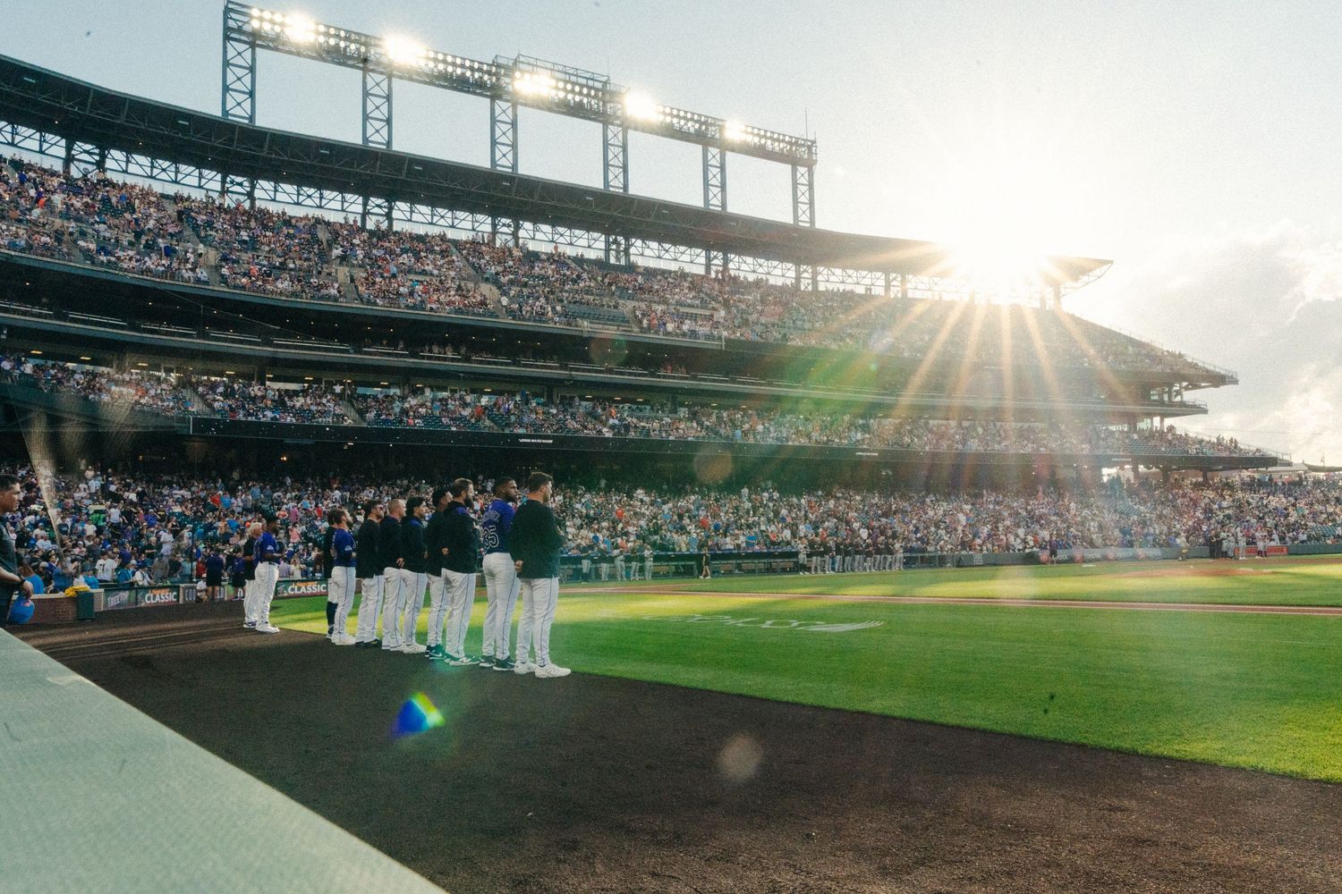 Baseball fans fill the stands at a sunlit stadium during a game as golden rays stream through the outfield bleachers.