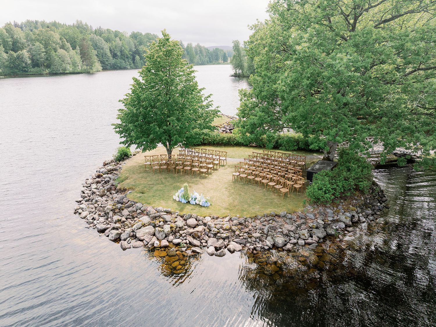 A panoramic view of a tranquil lakeside park with wooden benches and stone walls surrounded by lush green trees.