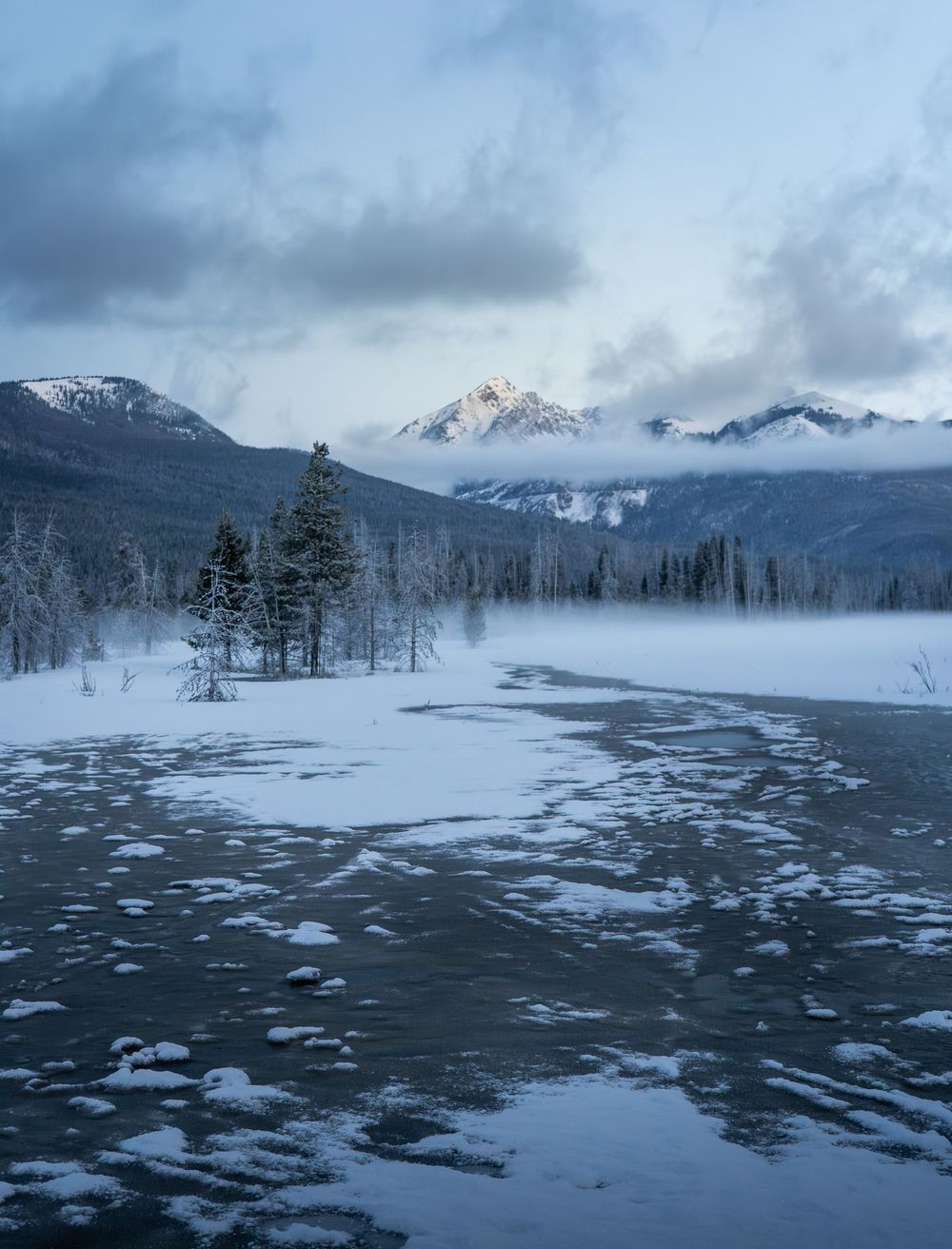 Snow-covered landscape with partially frozen lake reflects moody winter skies and mountain peaks in the background.