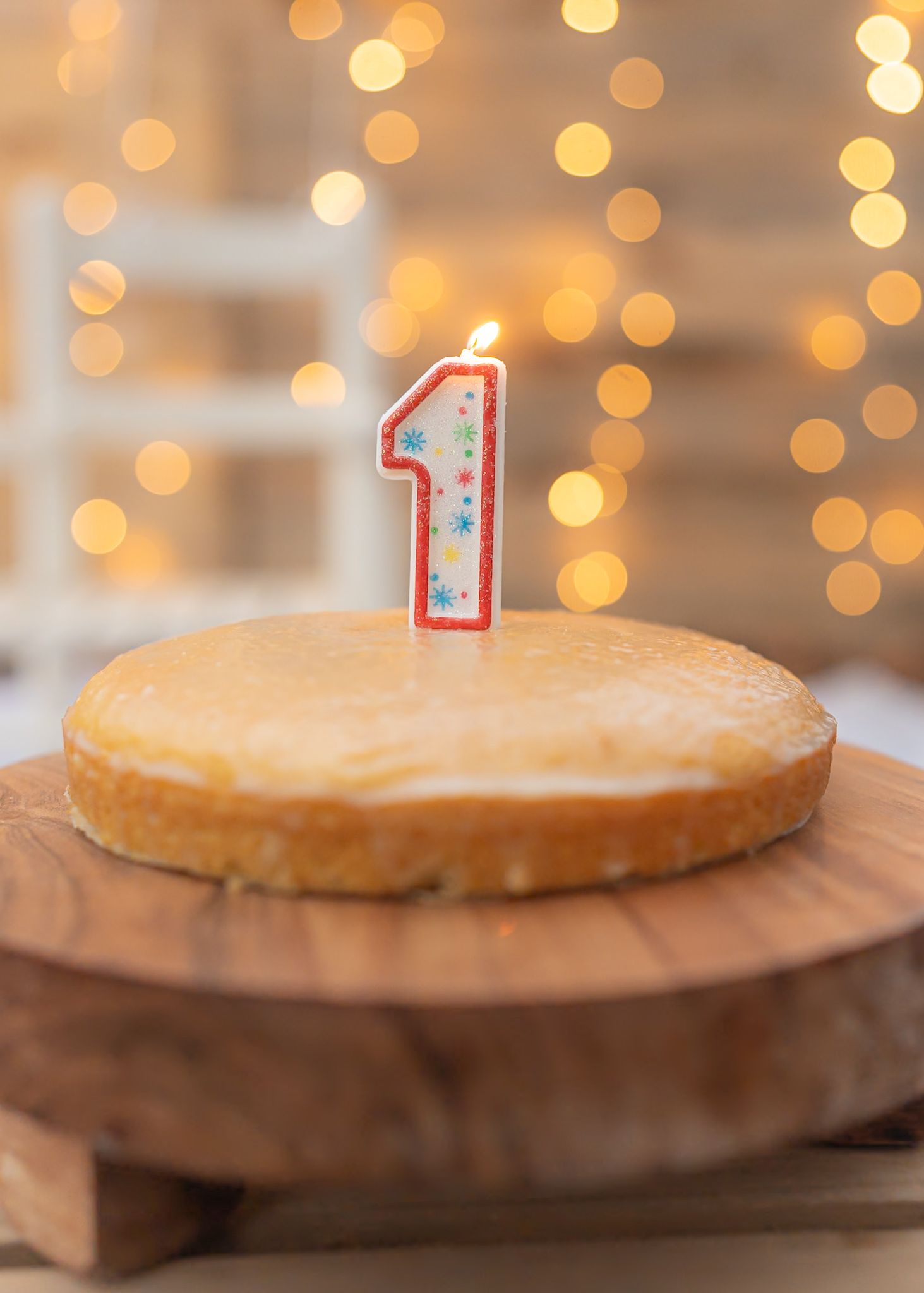 Round birthday cake with lit number one candle on wooden pedestal with bokeh lights.