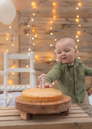 Baby in green shirt reaching for first birthday cake with twinkle lights in background.