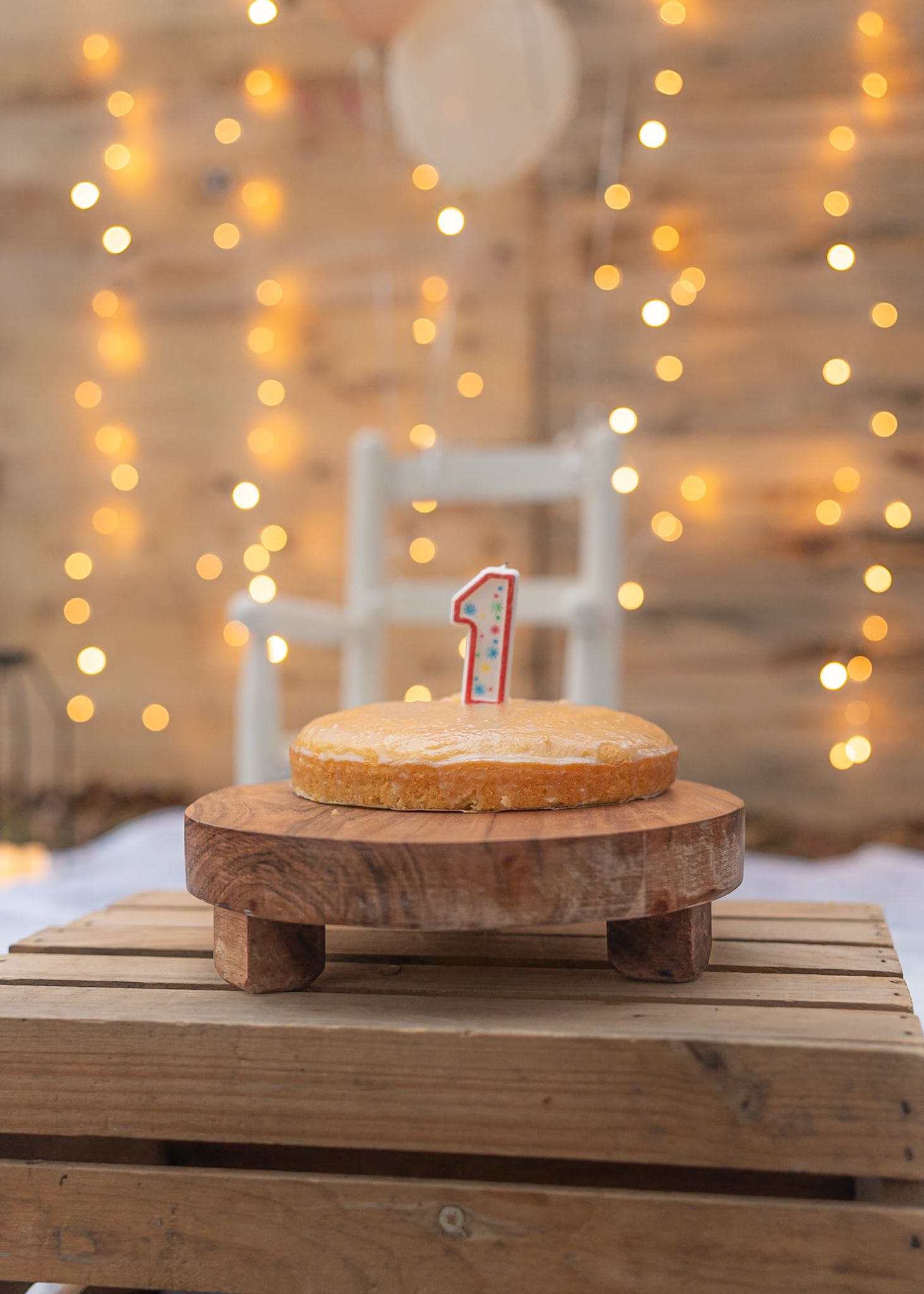 First birthday cake with number one candle on wooden stand against twinkle lights background.