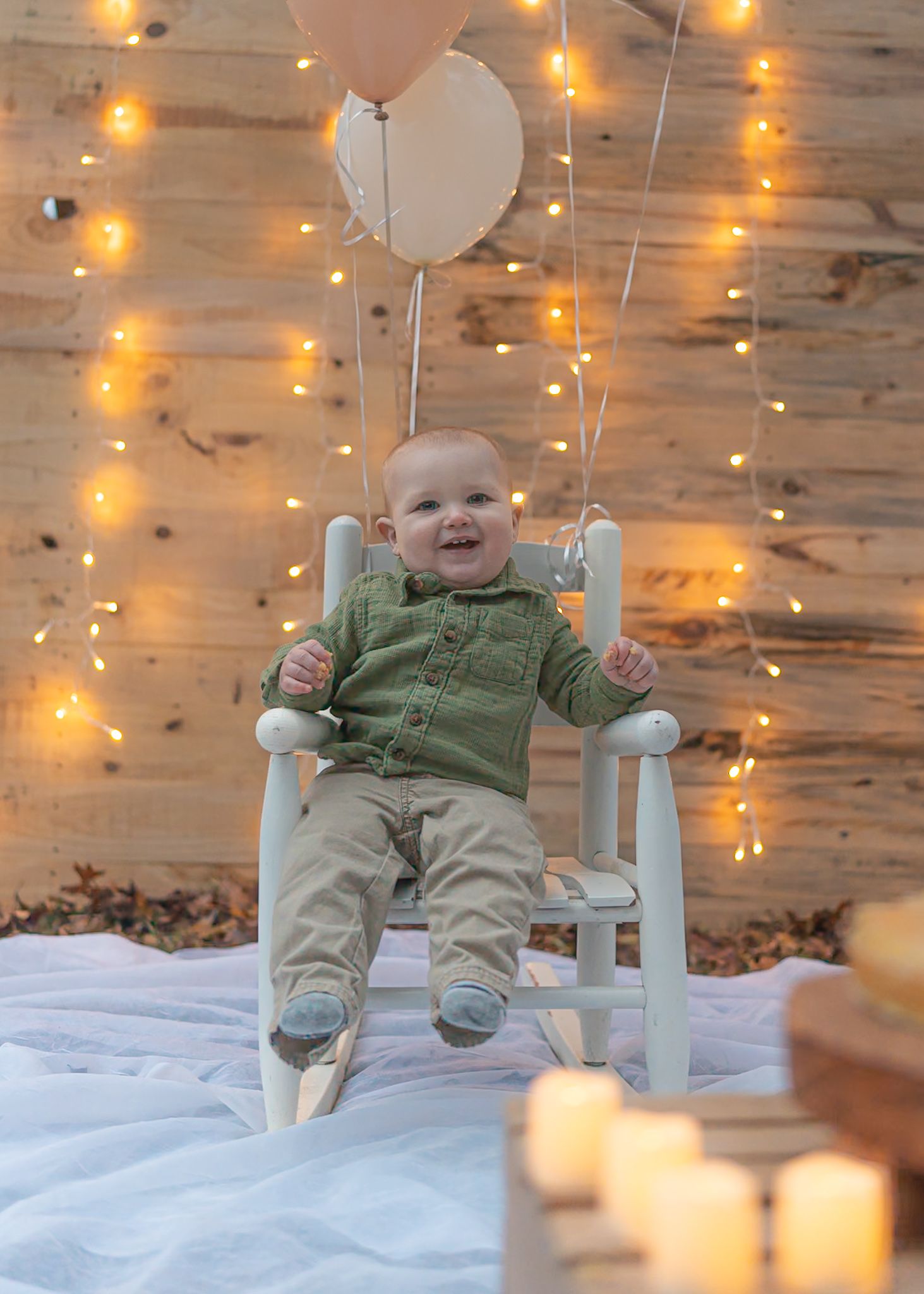 A baby sits in a light blue chair against a snowy backdrop with string lights overhead.