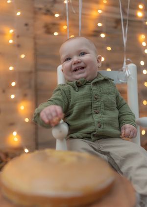 Festive first birthday scene with cake and white rocking chair against twinkle lights.