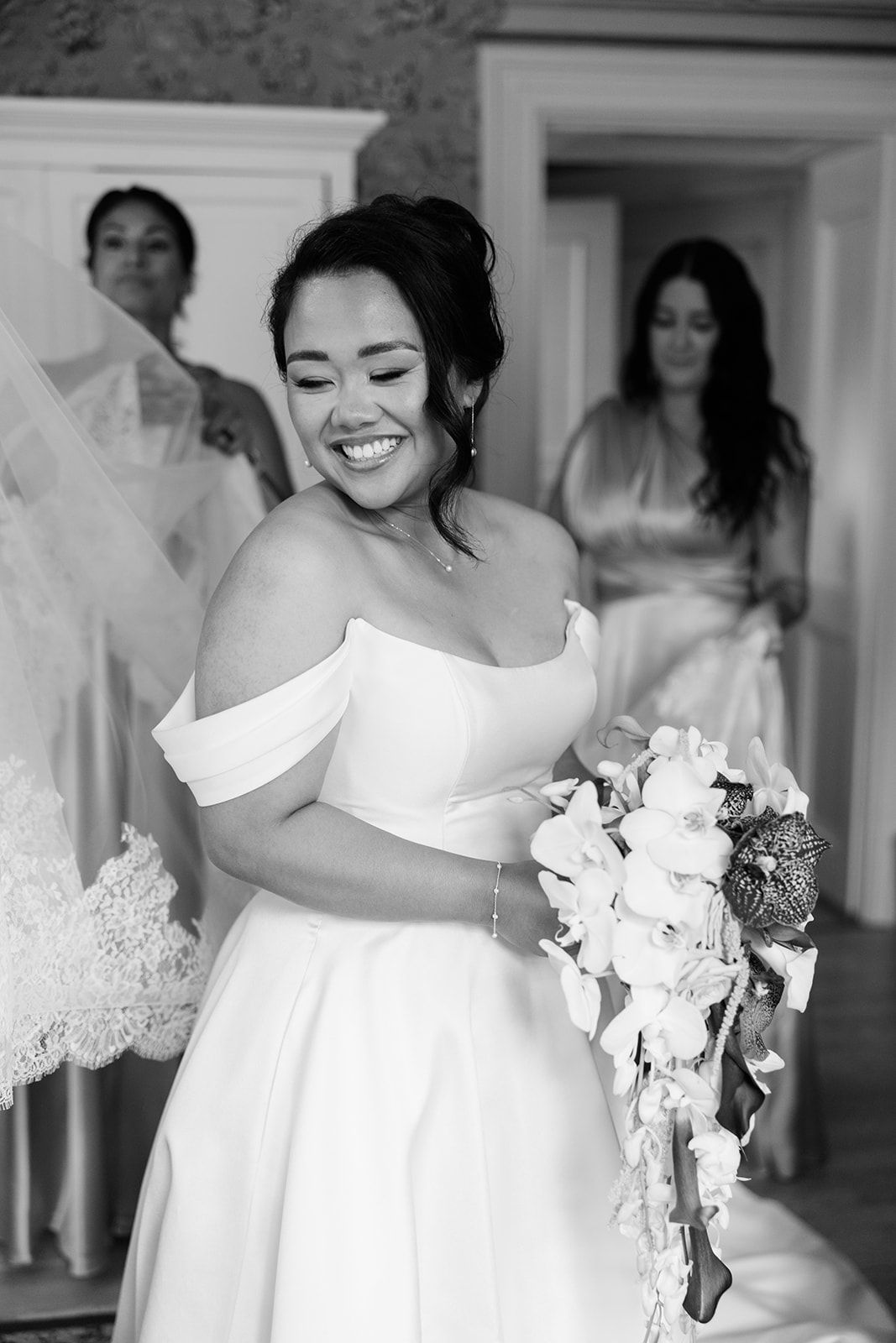 A joyful bride in an off-shoulder white wedding dress smiles while holding a bouquet of white flowers.