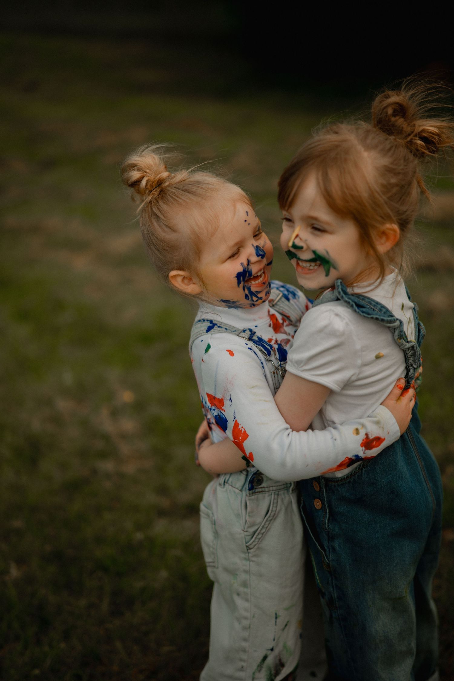 Two paint-covered toddler girls hugging and laughing outdoors, wearing denim overalls with colorful paint on faces.