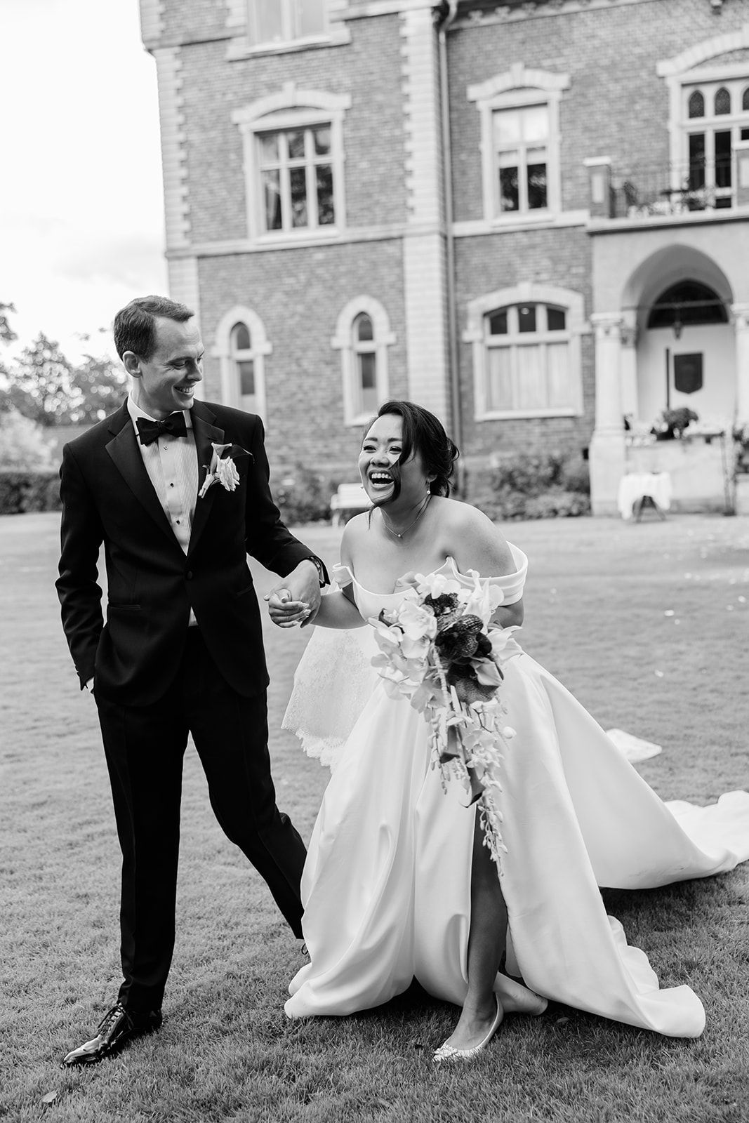 A joyful wedding couple shares candid moments together on a grassy lawn in front of a brick building.