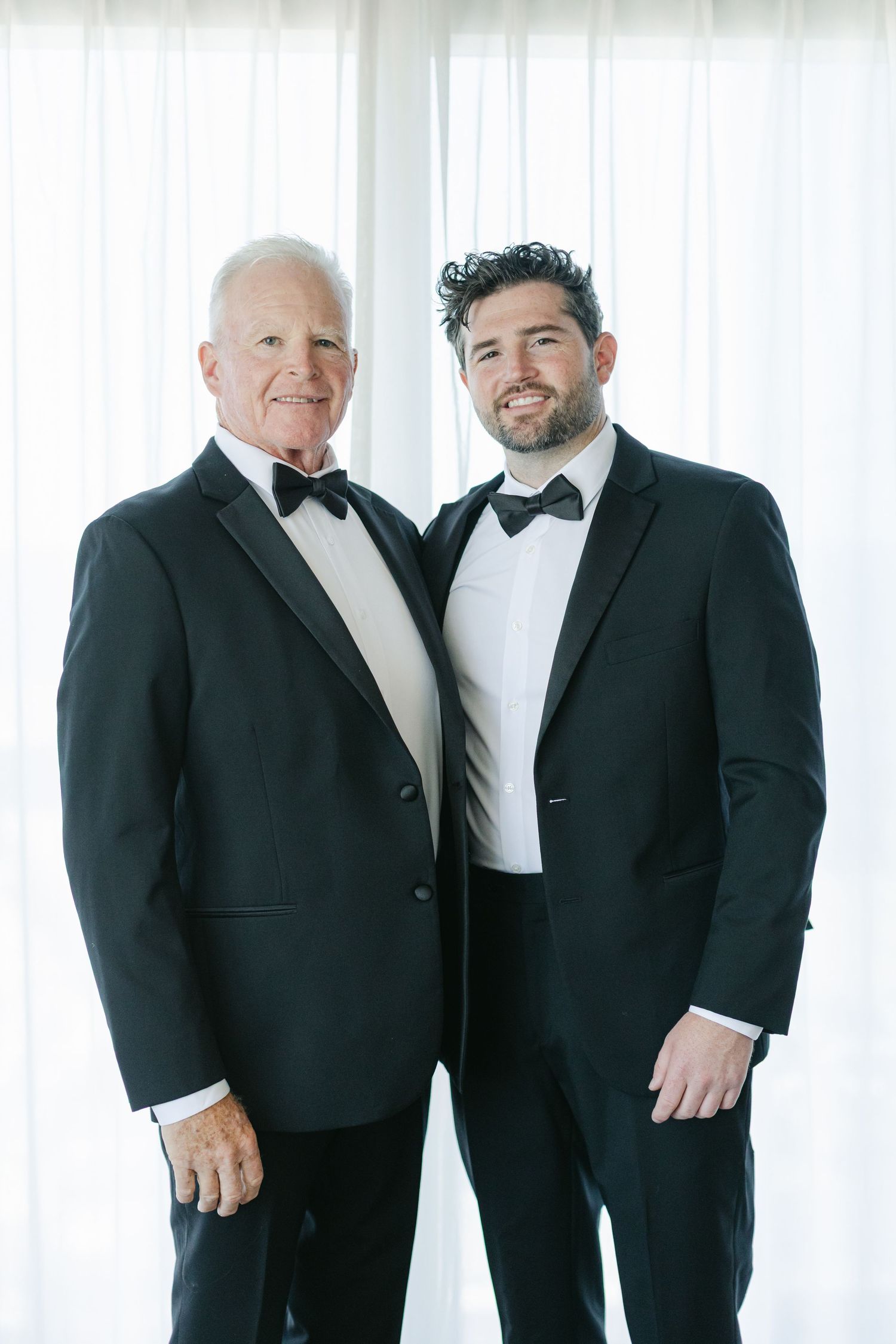 Two men in formal black and white tuxedos stand together smiling in front of a white backdrop.