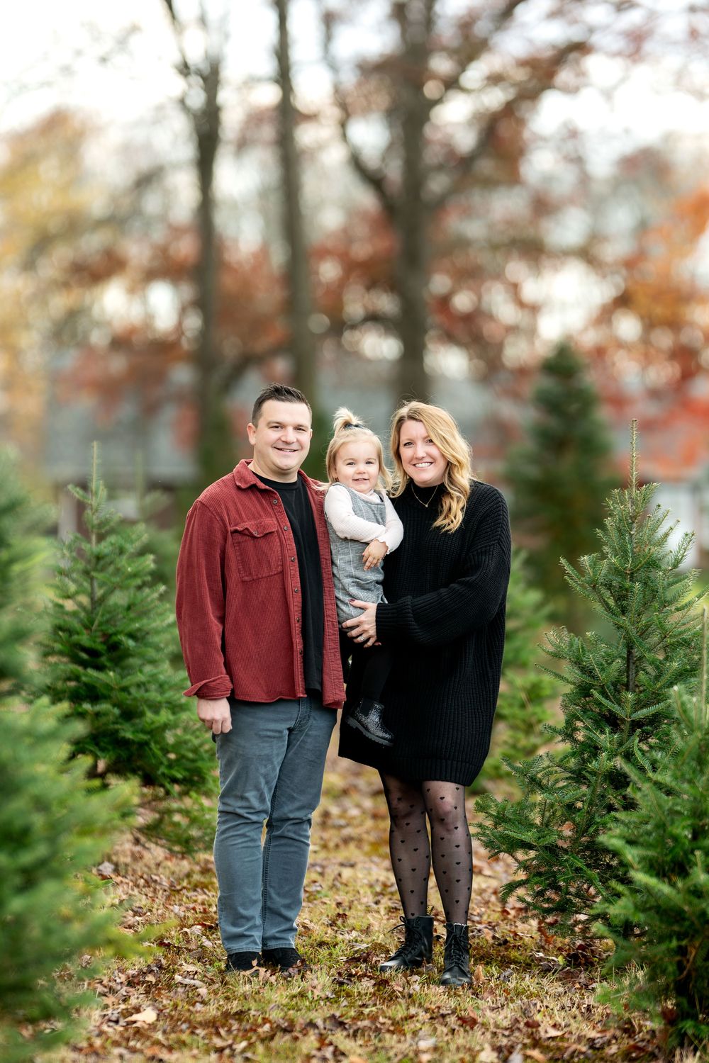 Happy family of three poses between rows of green Christmas trees at a tree farm.