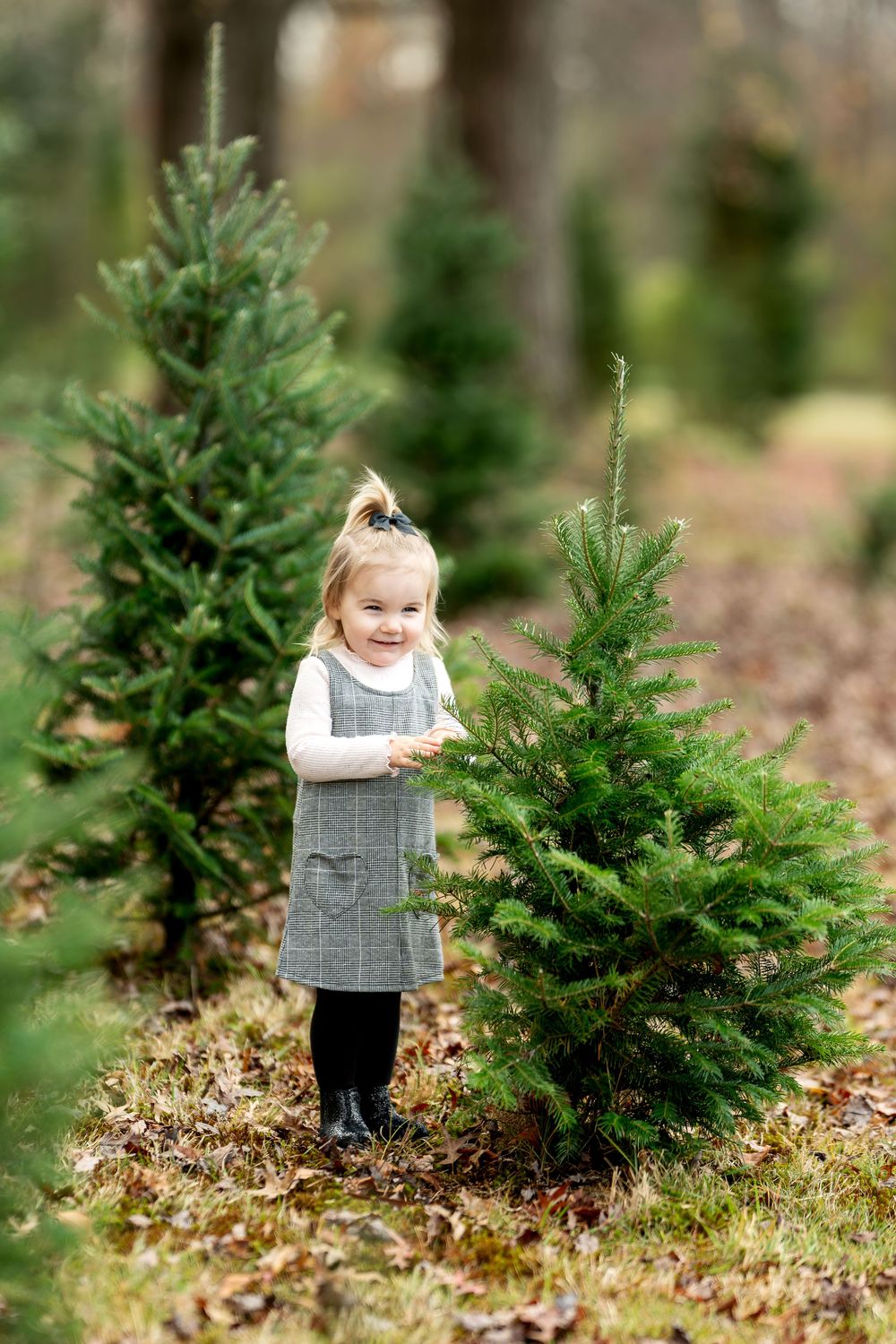 Smiling toddler girl in gray pinafore dress stands next to small evergreen trees outdoors.