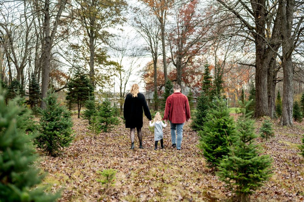 A family of three walks hand-in-hand through rows of Christmas trees on a fall day.