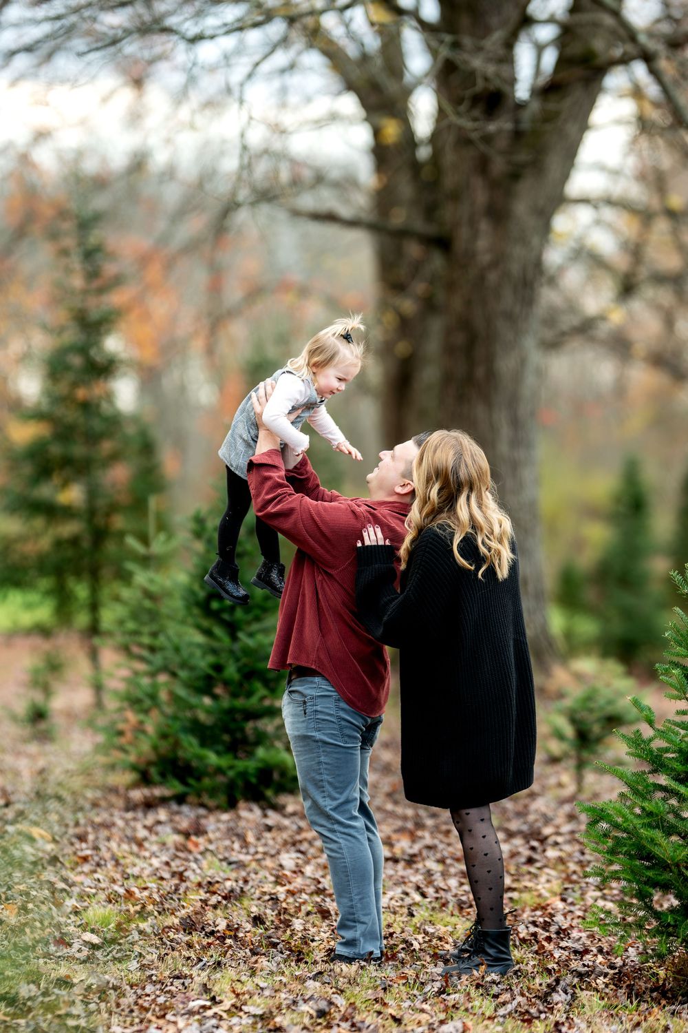 A mother lifts her toddler joyfully while a woman in black watches at a Christmas tree farm in autumn.
