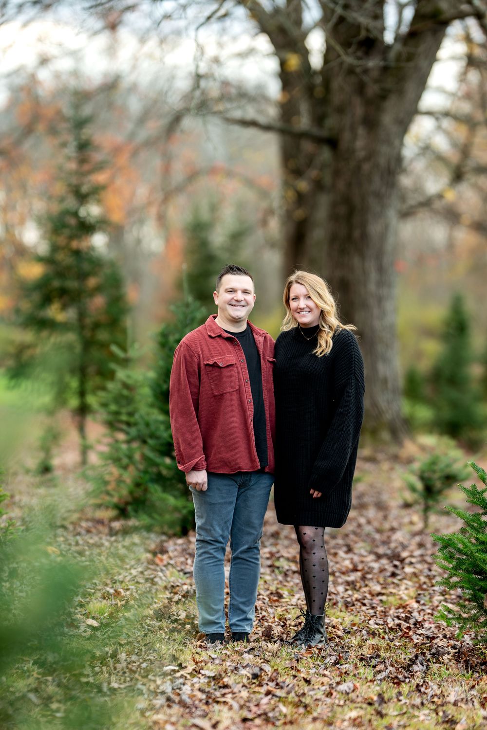 A smiling couple poses together among evergreen trees at an outdoor Christmas tree farm.