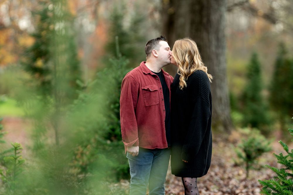 A couple shares a kiss among evergreen trees at an outdoor Christmas tree farm in fall.