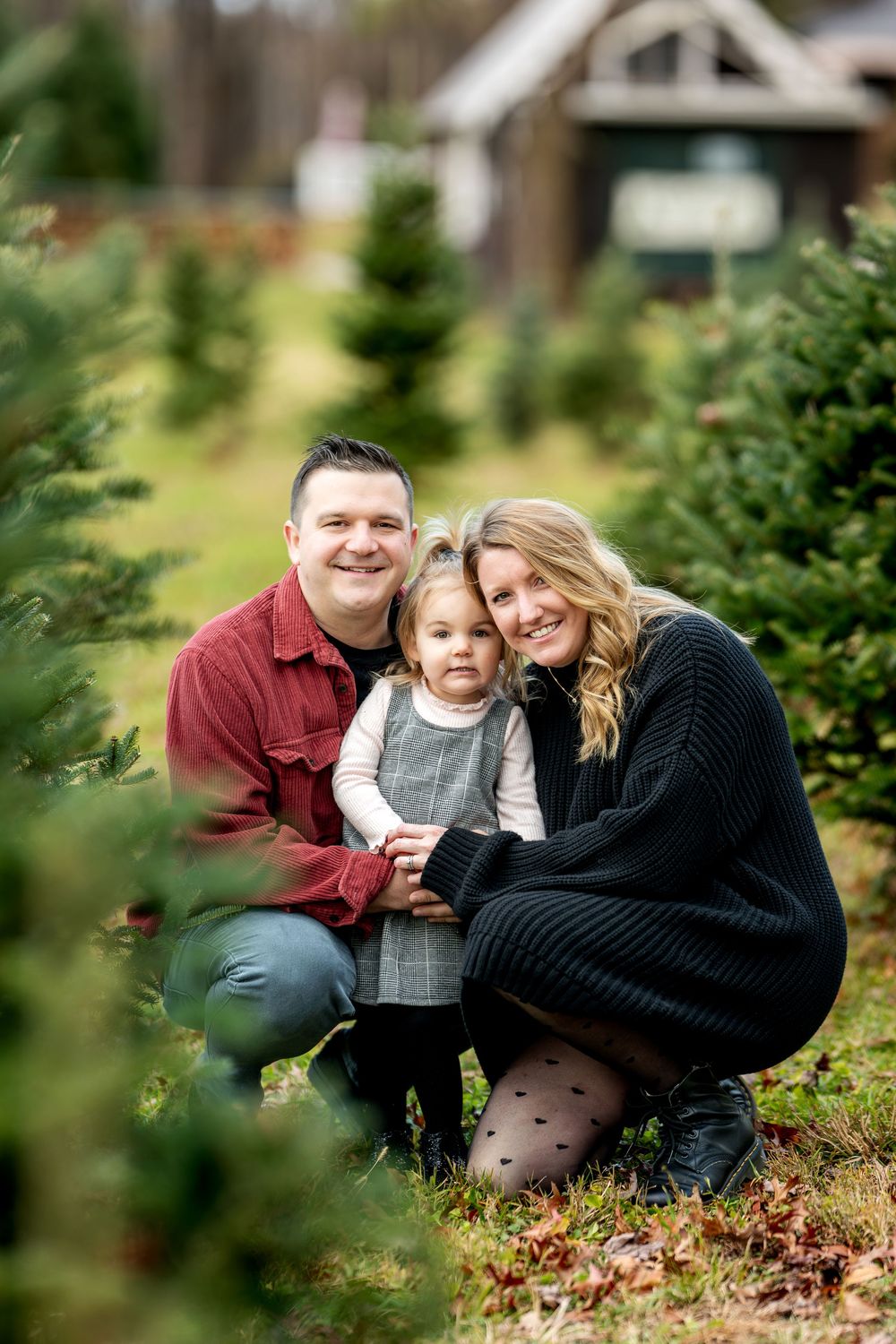 A smiling family of three poses together among Christmas trees at an outdoor tree farm.