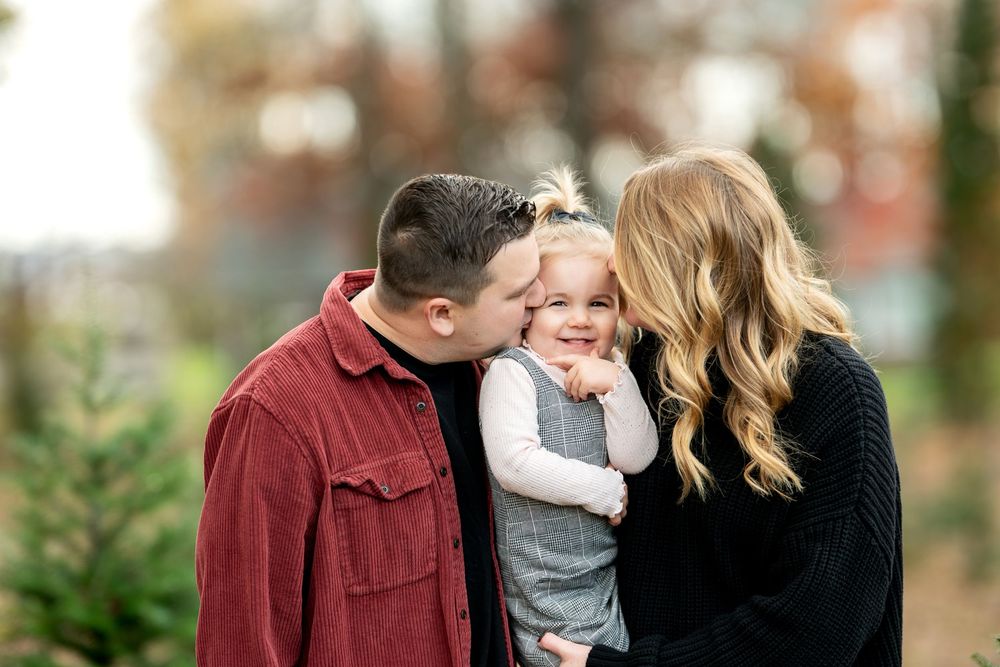Parents in red and black outfits kiss their smiling toddler on the cheeks outdoors.