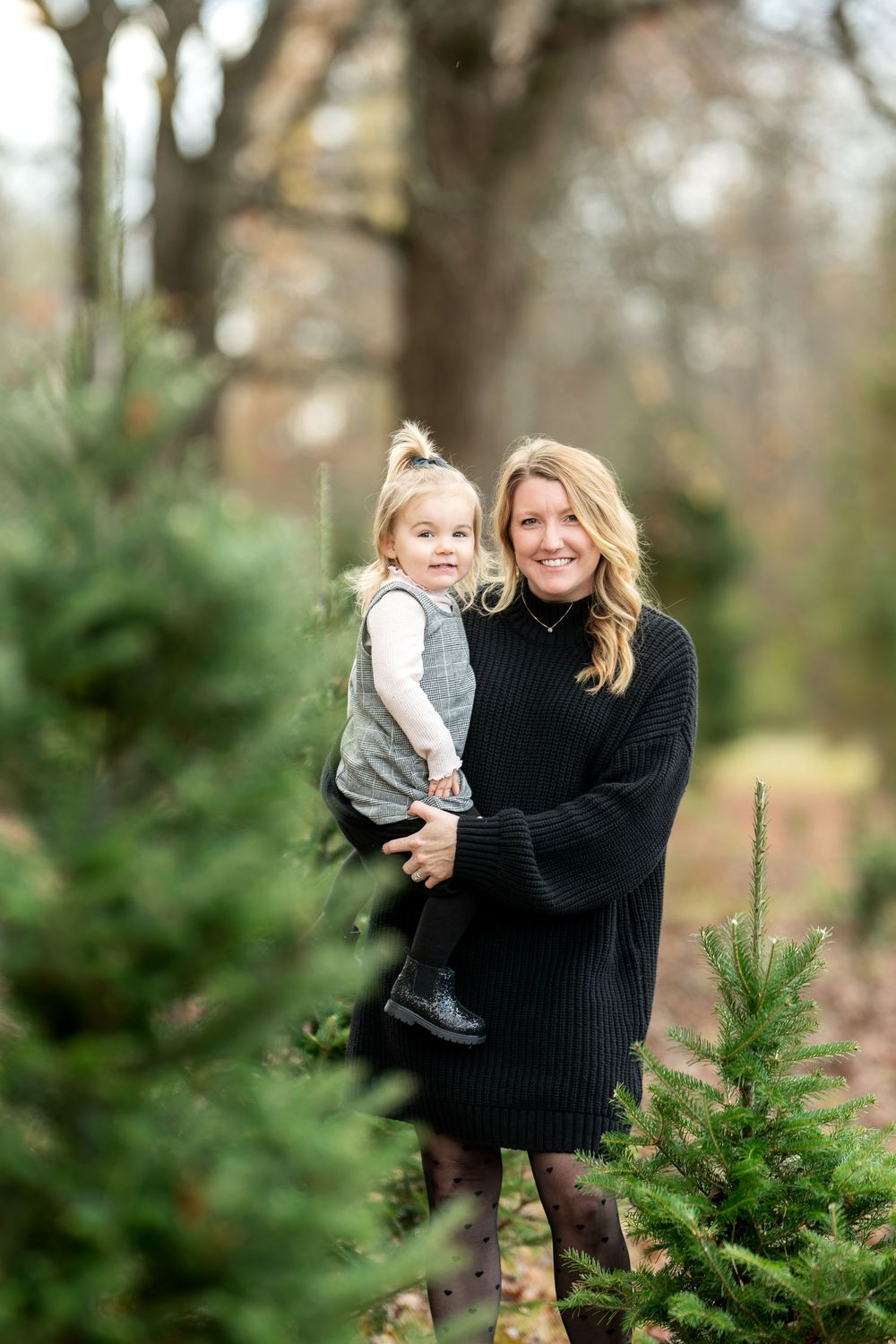 Woman in black sweater dress holds toddler girl among Christmas trees at a tree farm.