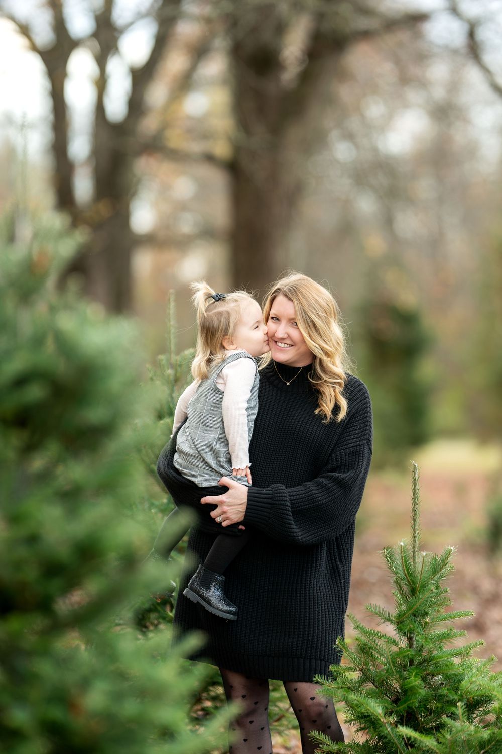 Mother in black dress cuddles smiling toddler daughter at an outdoor Christmas tree farm.