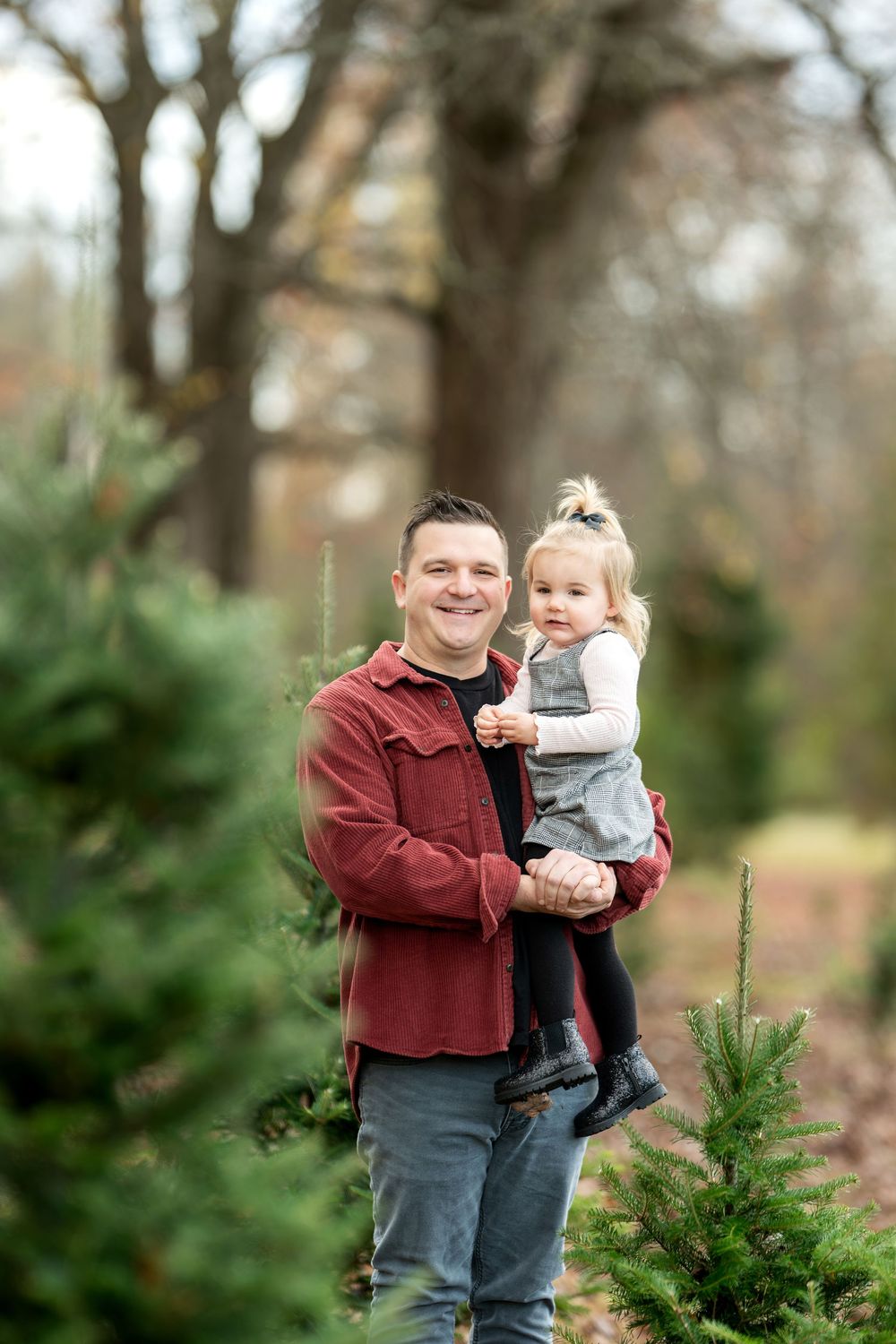 Smiling father in red shirt holds young blonde toddler daughter at a Christmas tree farm.