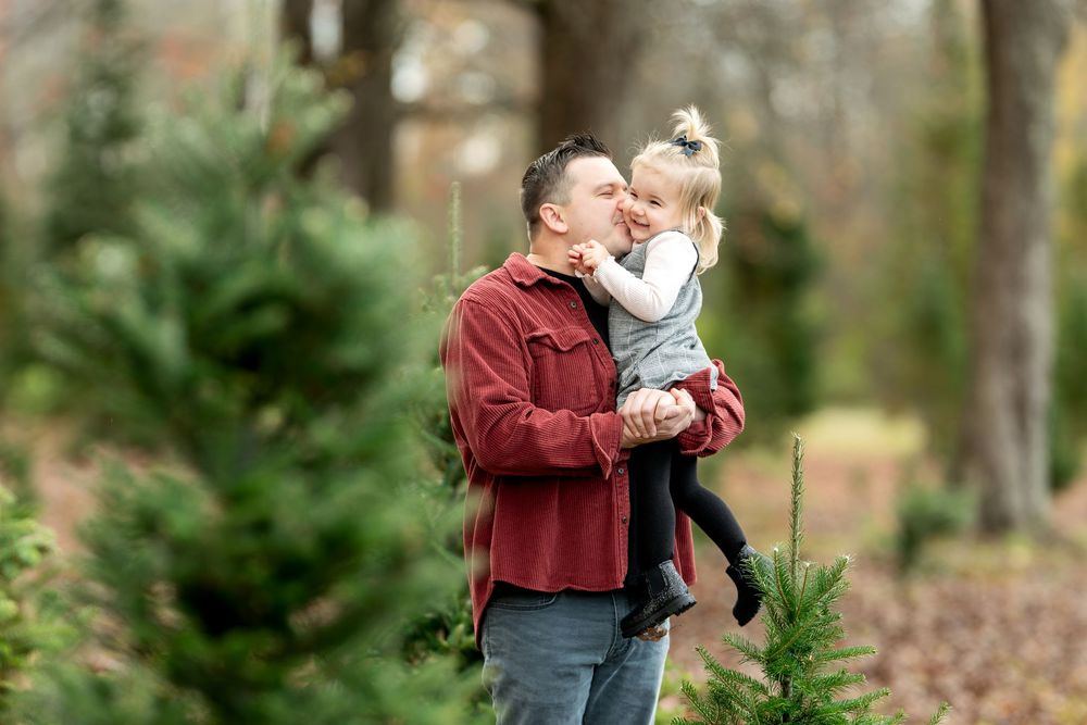 Dad in red shirt lifts and hugs his laughing toddler girl among Christmas trees outdoors.