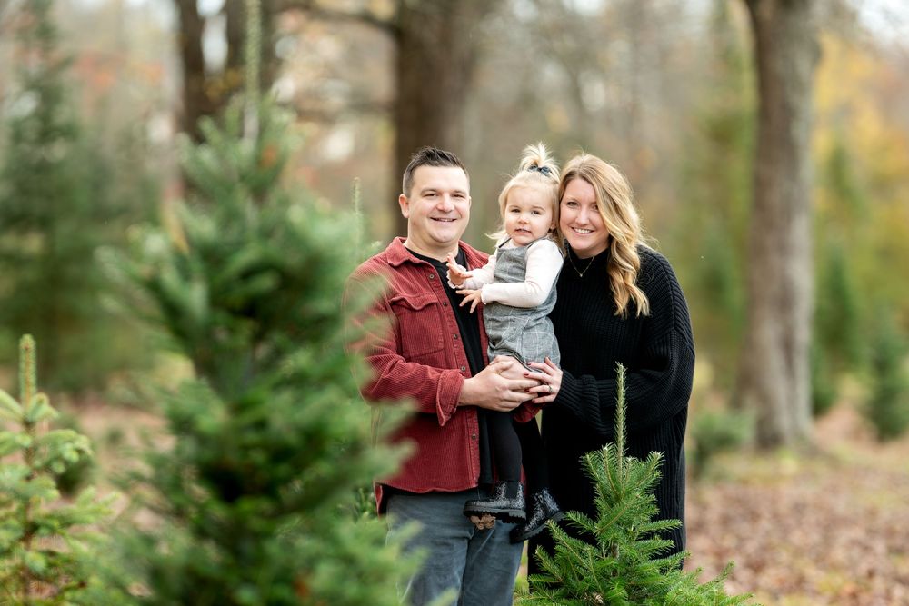 Smiling family with toddler poses together between evergreen trees at a Christmas tree farm.