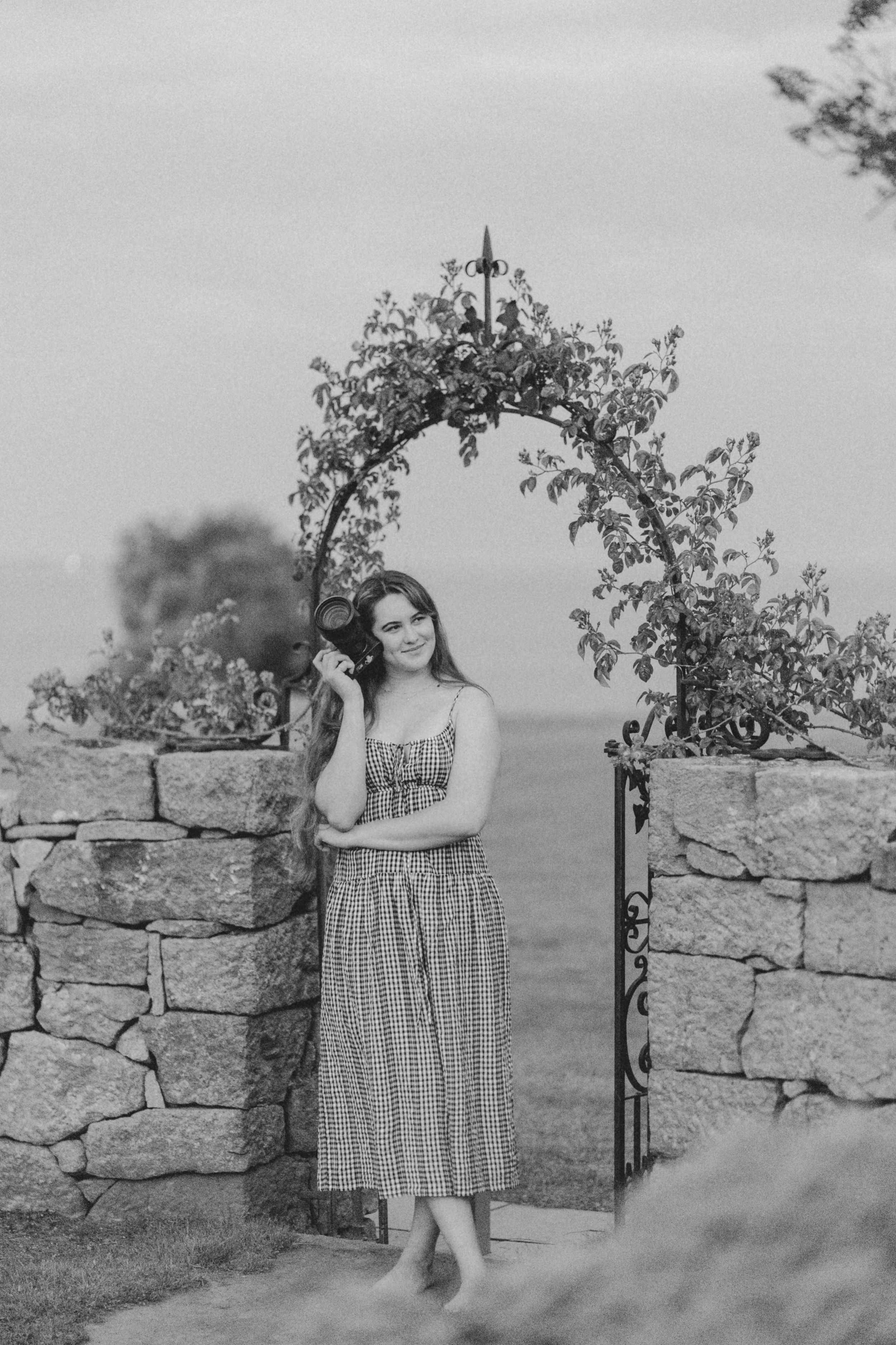 A black and white photo series of someone in a gingham dress posing against a rustic stone wall.