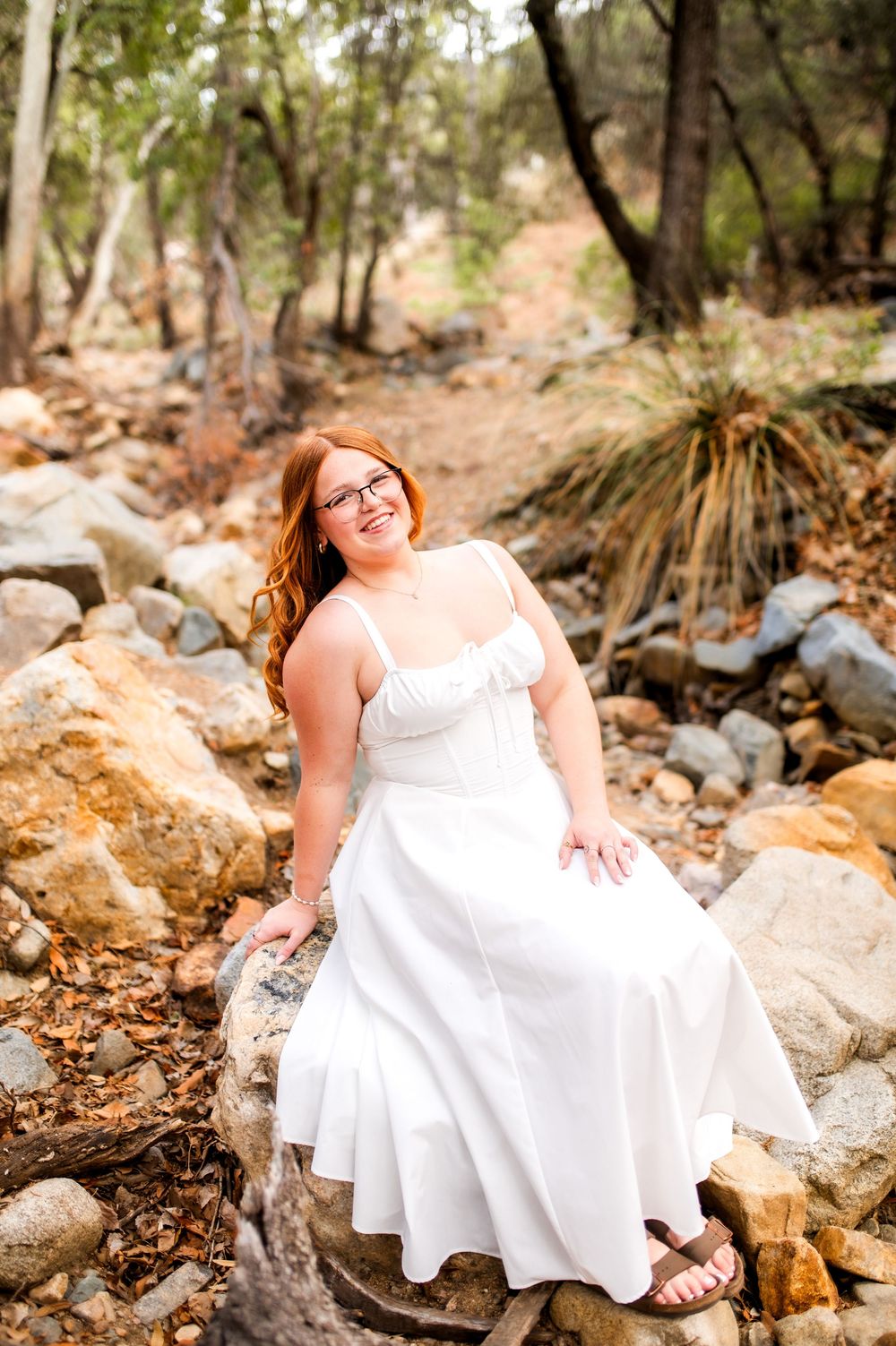 Smiling redhead woman in white dress sitting on rocks in a wooded outdoor setting.