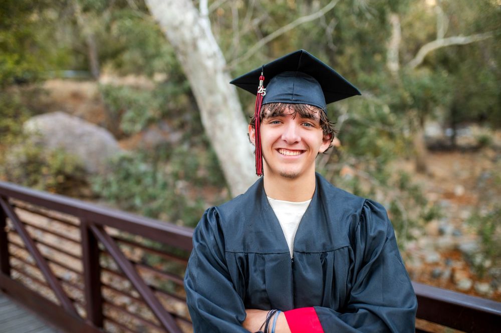 Smiling young man in graduation cap and gown standing on a wooden bridge.