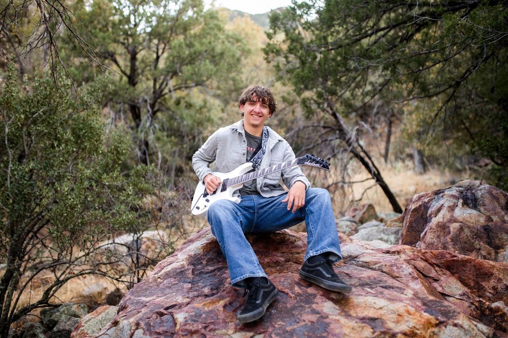 Young man sitting on a red rock playing a white electric guitar outdoors.