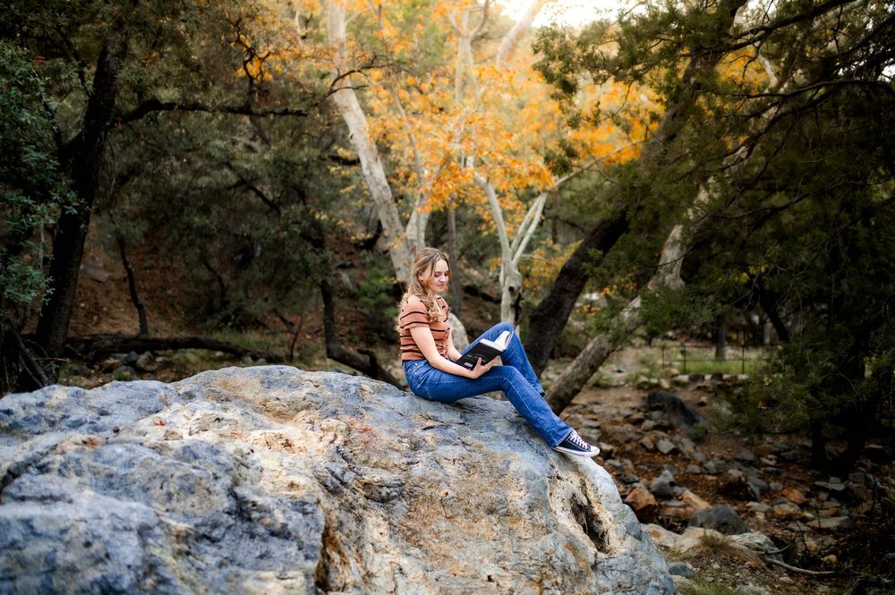 A young woman sits on a large boulder reading a book surrounded by colorful autumn foliage.