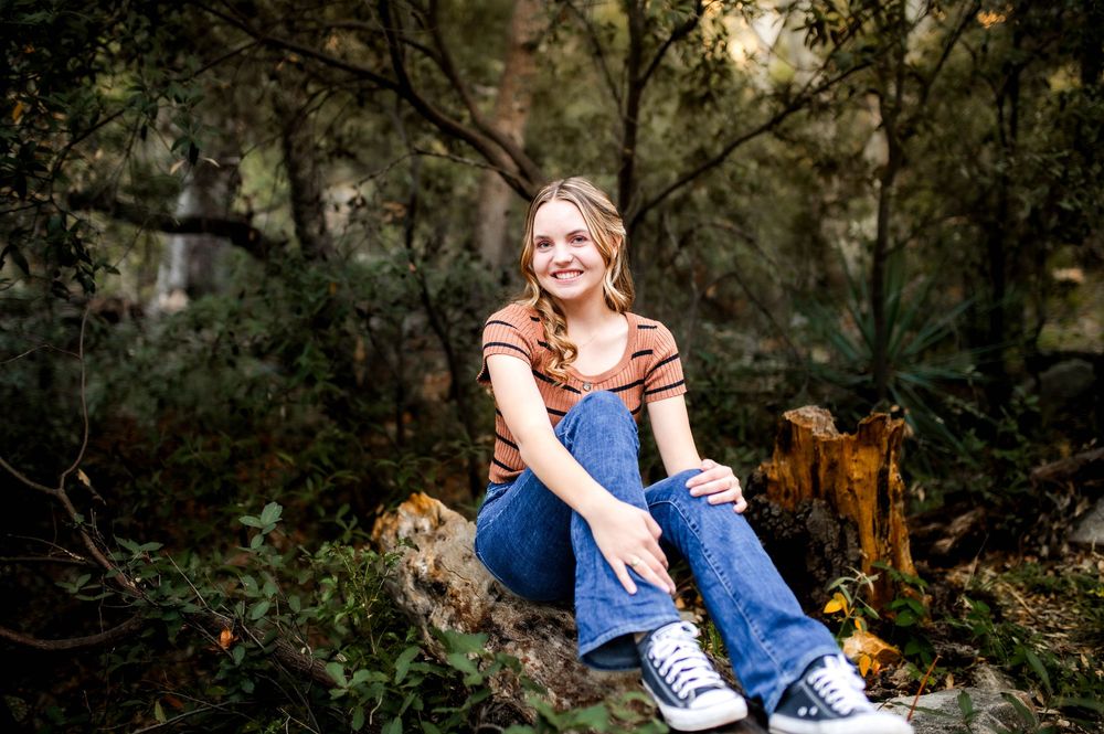 A smiling teenage girl in a rust top and jeans sits on a log in a lush green woodland setting.